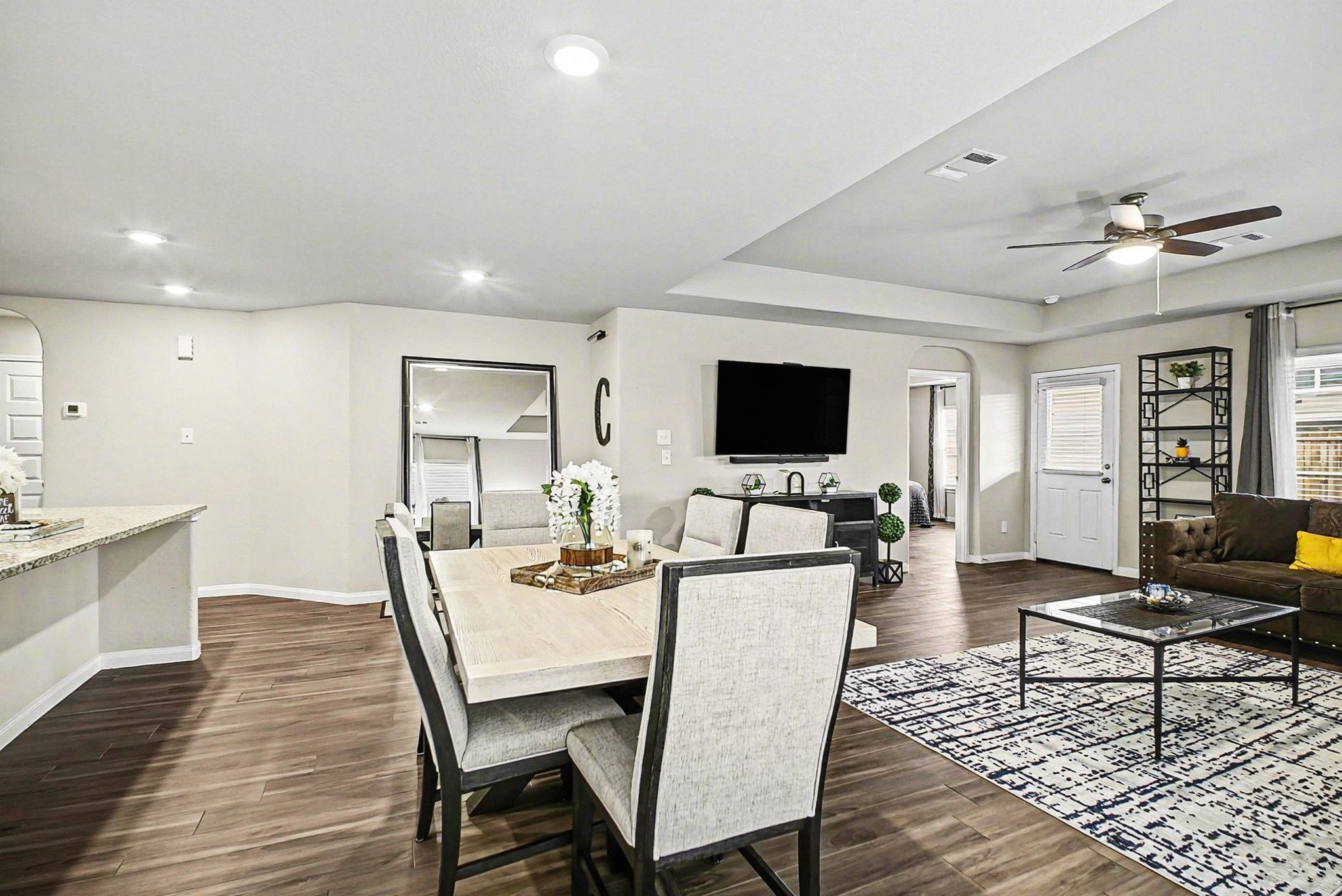 4906 Luke Matthew Drive Rosharon, TX 77583 - Photo 9 of 34 This home features a modern open-concept living and dining area with wood flooring. It features a dining table, comfortable seating, a wall-mounted TV, and a ceiling fan. The space has neutral tones and ample natural light, with a view of a kitchen counter and access to additional rooms.