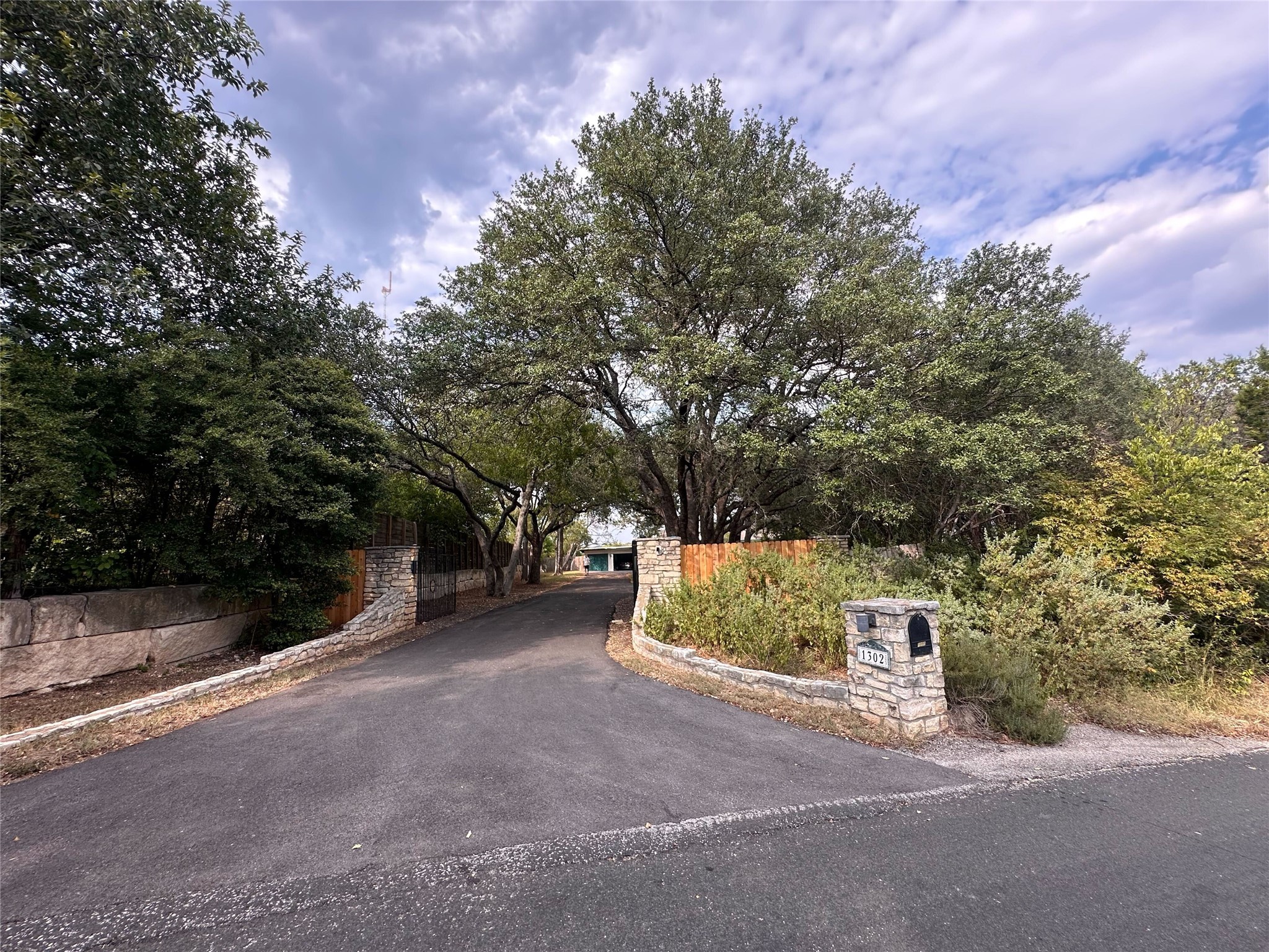 1302 Ridgecrest Drive Austin, TX 78746 - Photo 2 of 19 Mature trees arch over a paved driveway leading past stone retaining walls.