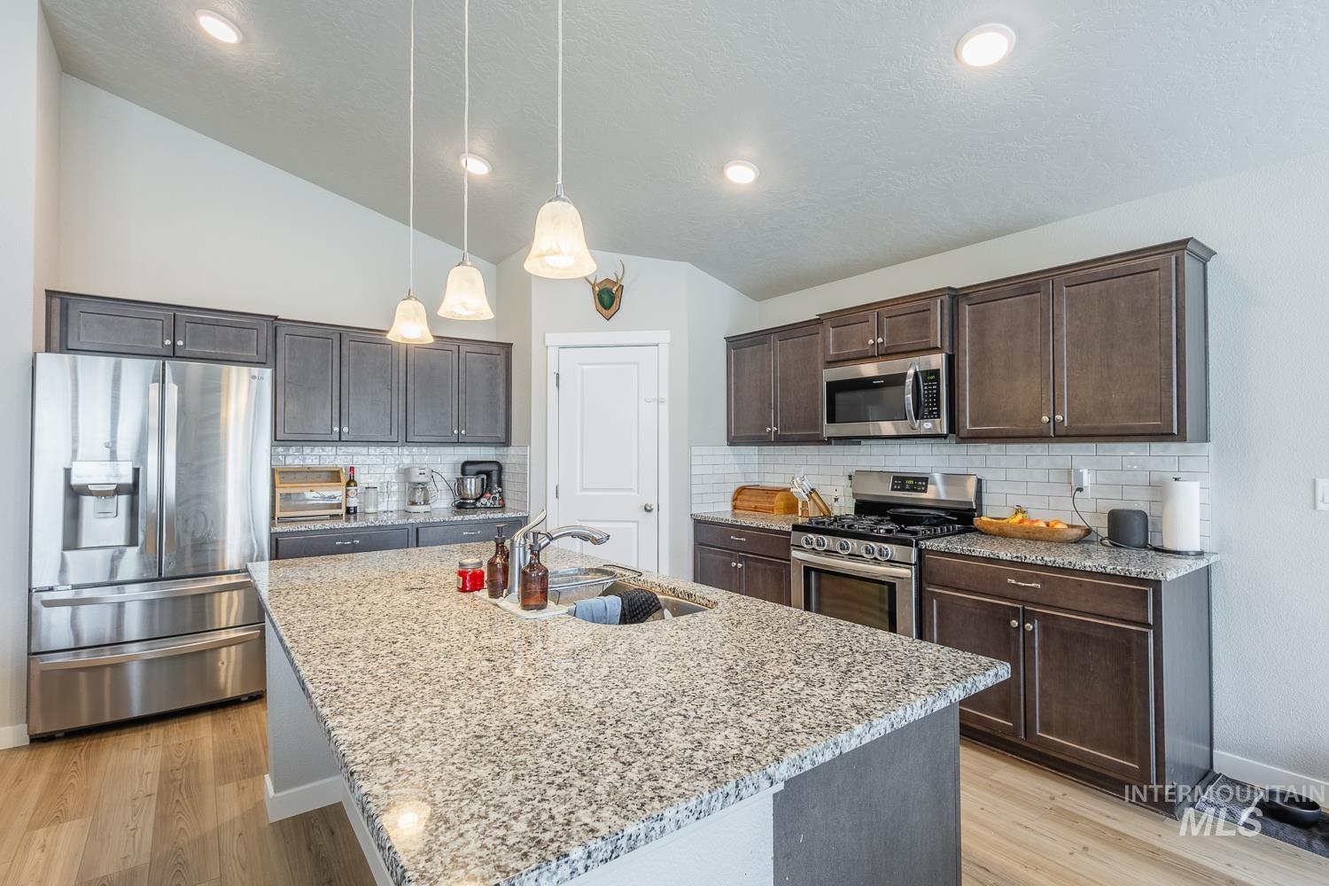12623 Shadow Peak Street Caldwell, ID 83607 - Photo 14 of 50 Contemporary kitchen featuring a large island with a surface-mounted sink, light-toned granite countertops, dark wood cabinetry, stainless steel appliances, and wood-finish flooring
