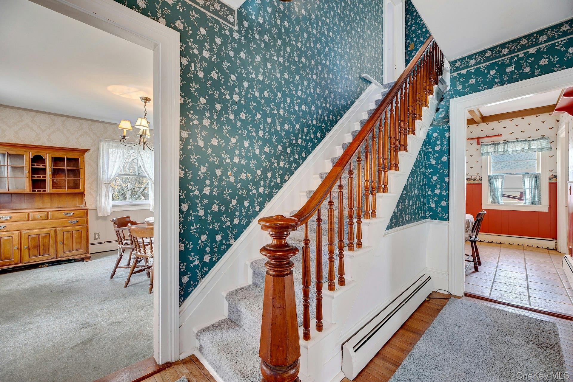 1760 Albertson Lane Greenport, NY 11944 - Photo 3 of 40 Staircase with wallpapered walls, a baseboard heating unit, a chandelier, wood finished floors, and a wainscoted wall