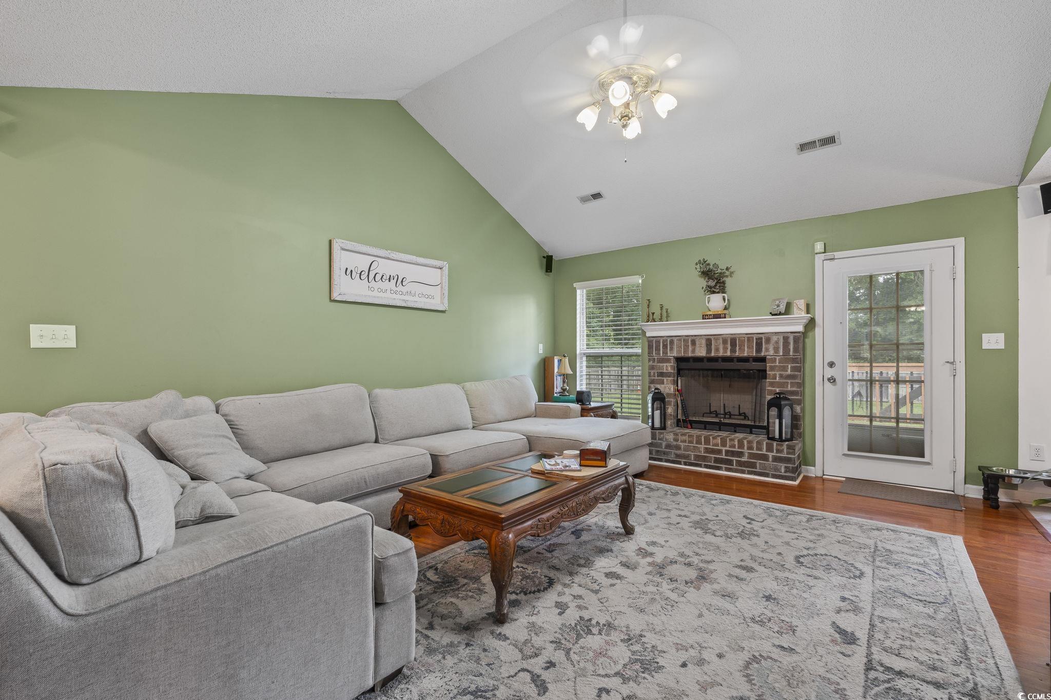 Living room with wood finished floors, vaulted ceiling, a brick fireplace, and a ceiling fan