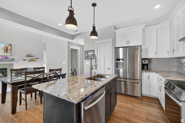 7872 Farm House Lane Midvale, UT 84047 - Photo 14 of 39 Modern kitchen featuring an island with a double sink, stainless steel appliances, white upper cabinetry, gray subway tile backsplash, and wood-finish flooring