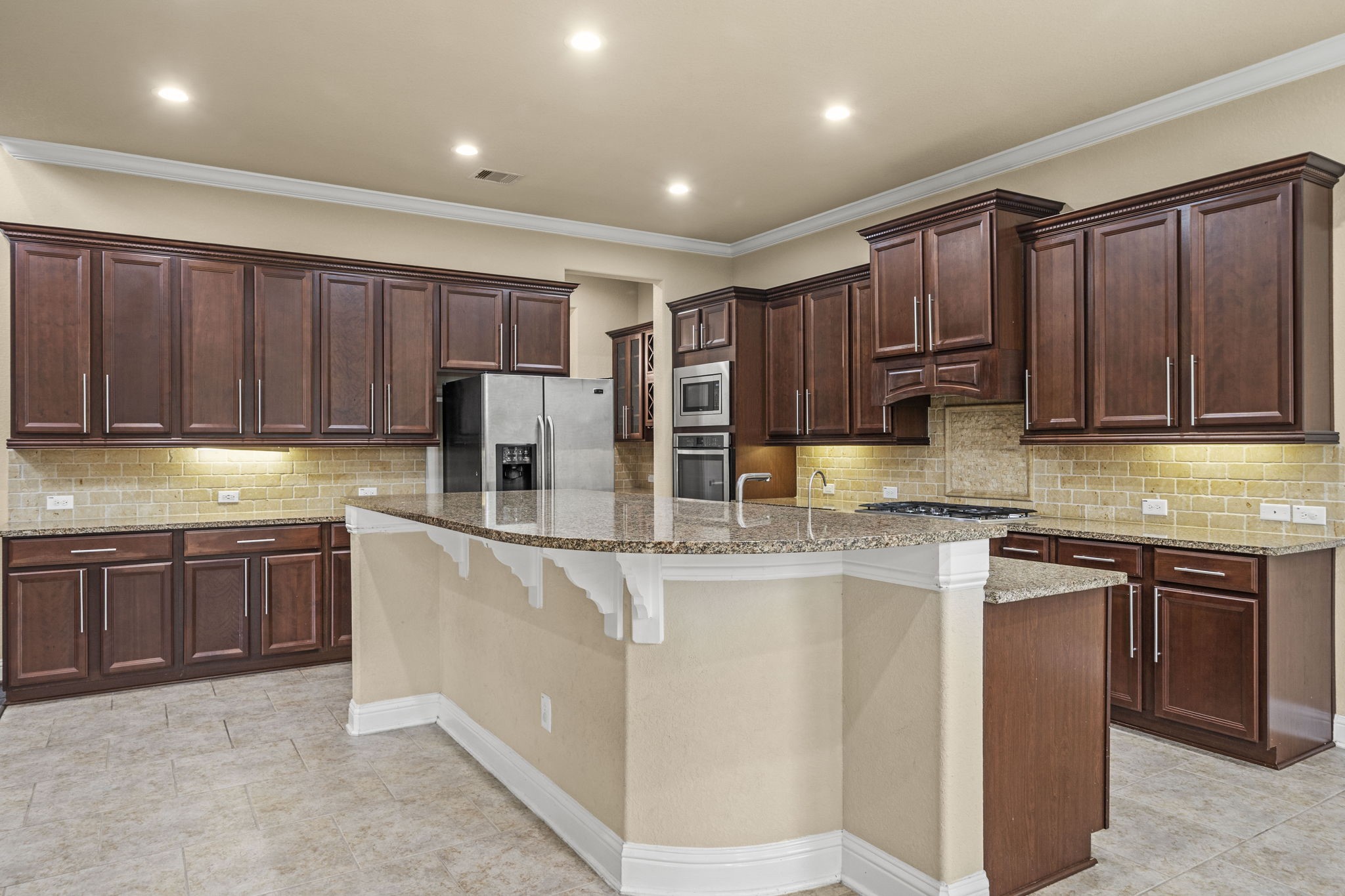 14215 Spindle Arbor Road Cypress, TX 77429 - Photo 14 of 47 This spacious kitchen features elegant dark wood cabinetry, a large granite-topped island with a sink, and stainless steel appliances. The neutral tile flooring and warm lighting create a welcoming atmosphere ideal for cooking and entertaining.
