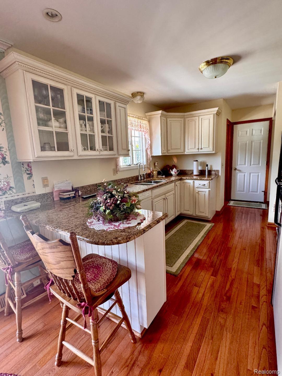 6686 South Schott Road Mayville, MI 48744 - Photo 3 of 25 Kitchen featuring wood-finish flooring, granite countertops, white cabinetry with glass-front uppers, a kitchen island with beadboard paneling, and a stainless steel sink