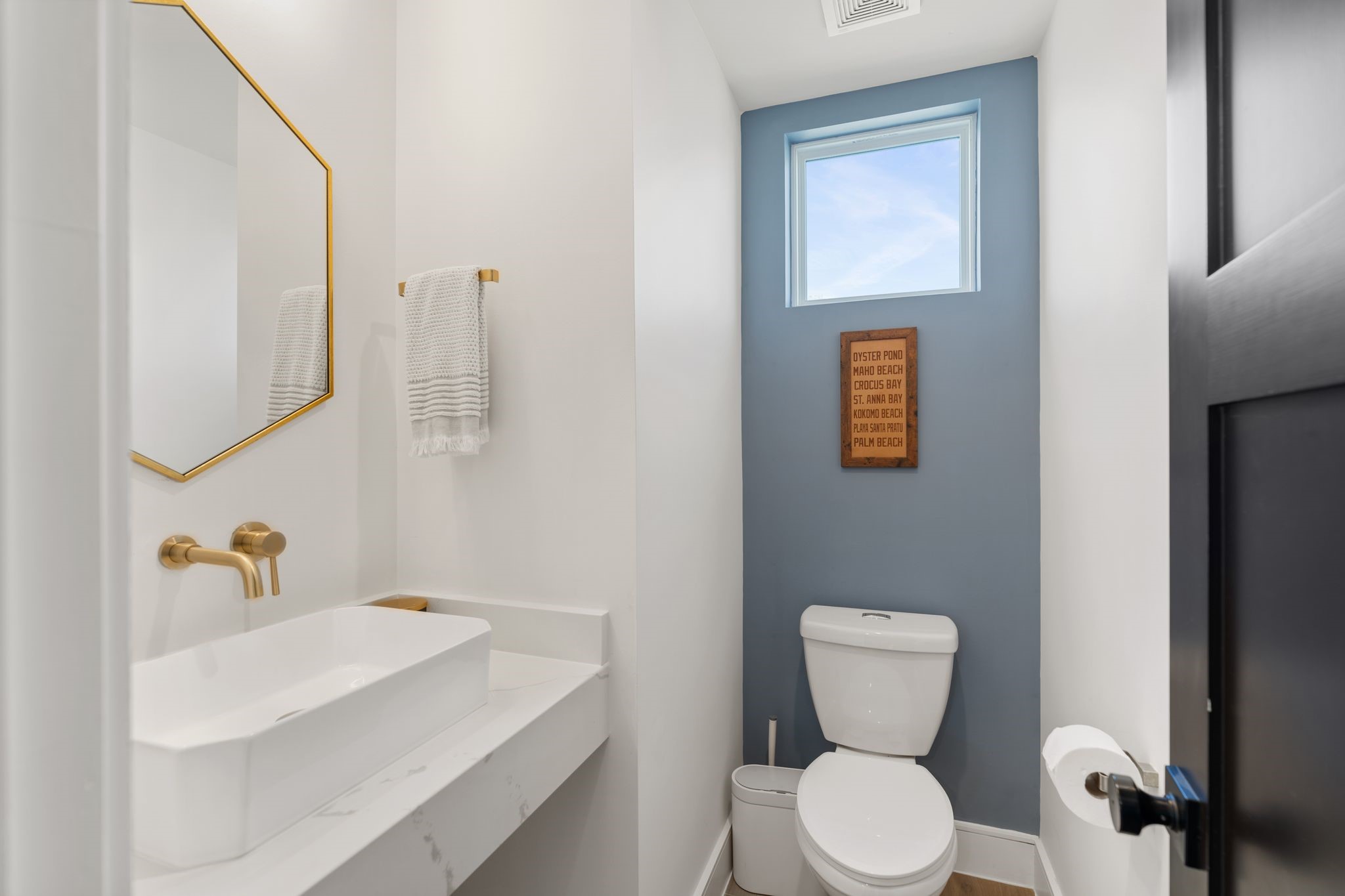 2626 Reagan Street Houston, TX 77009 - Photo 29 of 50 The upstairs powder bath features a sleek floating vanity with a vessel sink and marble countertop, paired with a wall-mounted gold faucet and a decorative gold framed mirror. The blue wall adds depth and character, while the window above allows natural light into the space, completing this thoughtfully designed bathroom with modern finishes and stylish details.