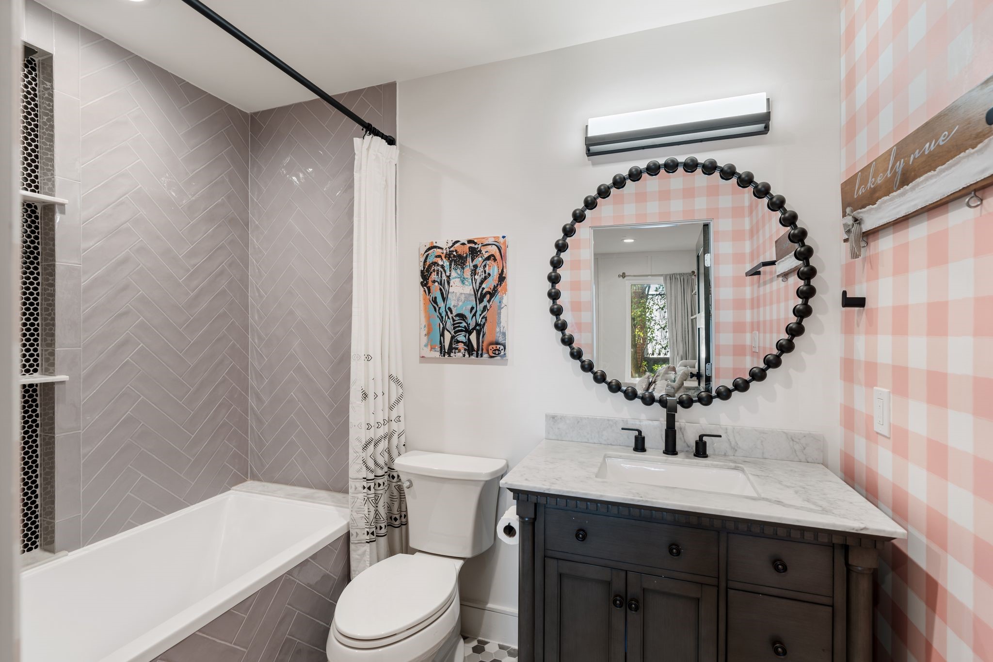 2626 Reagan Street Houston, TX 77009 - Photo 31 of 50 The en-suite bathroom features a marble countertop vanity with black fixtures, a decorative round mirror with bead detailing, and playful gingham wallpaper. The tub/shower combination is finished with floor-to-ceiling herringbone tile and built-in shower niches complemented by a hexagon tile flooring.