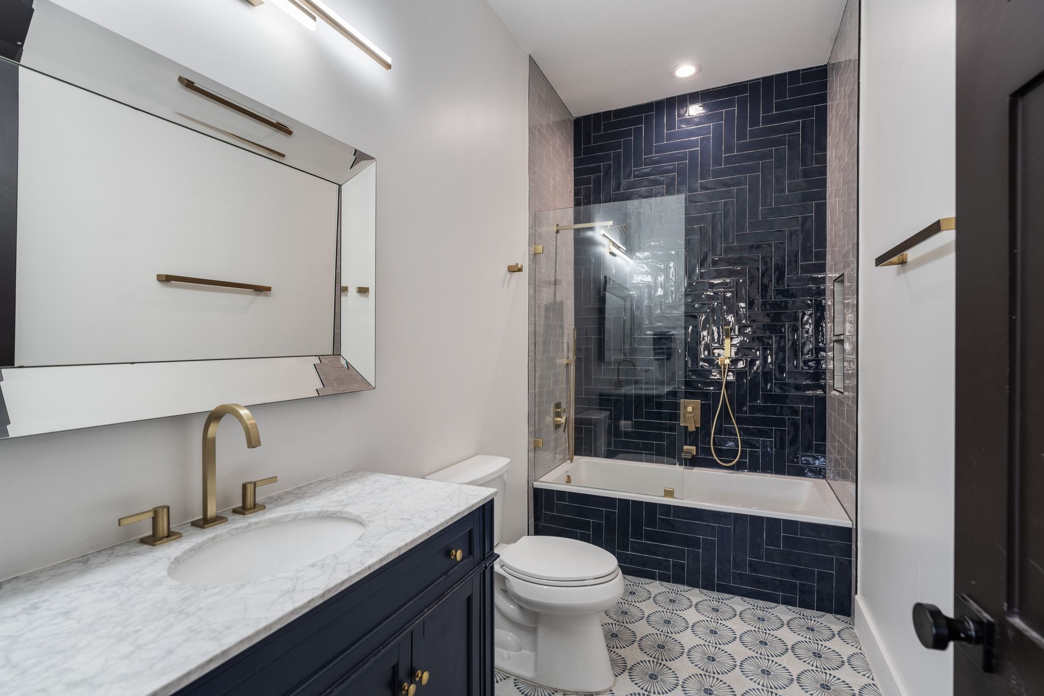 2626 Reagan Street Houston, TX 77009 - Photo 35 of 50 Gorgeous ensuite bath with a rich navy vanity topped with a marble countertop, paired with sleek gold hardware and a statement gold faucet. oversized decorative mirror and modern lighting keep the space bright, while the bold pattern tile flooring adds personality. The tub / shower is wrapped in dramatic dark blue herringbone tile with a glass splash panel and coordinating gold fixtures, creating a high-end, designer look from top to bottom.