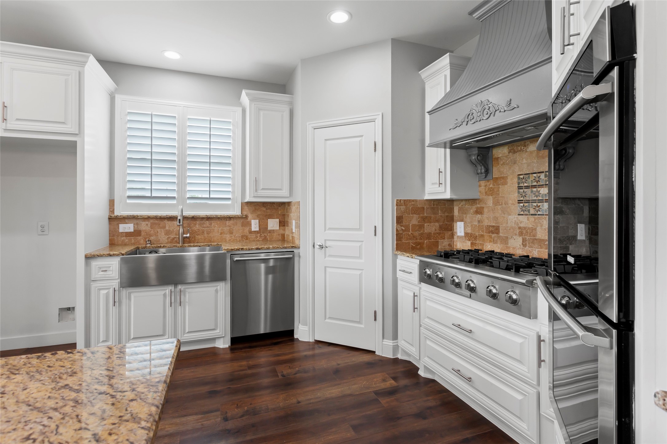 610 South Shady Lane La Porte, TX 77571 - Photo 15 of 50 This kitchen features elegant white cabinetry, a farmhouse sink, and stainless steel appliances, including a large gas range. The warm stone backsplash and rich laminate wood floors add a touch of luxury and warmth.