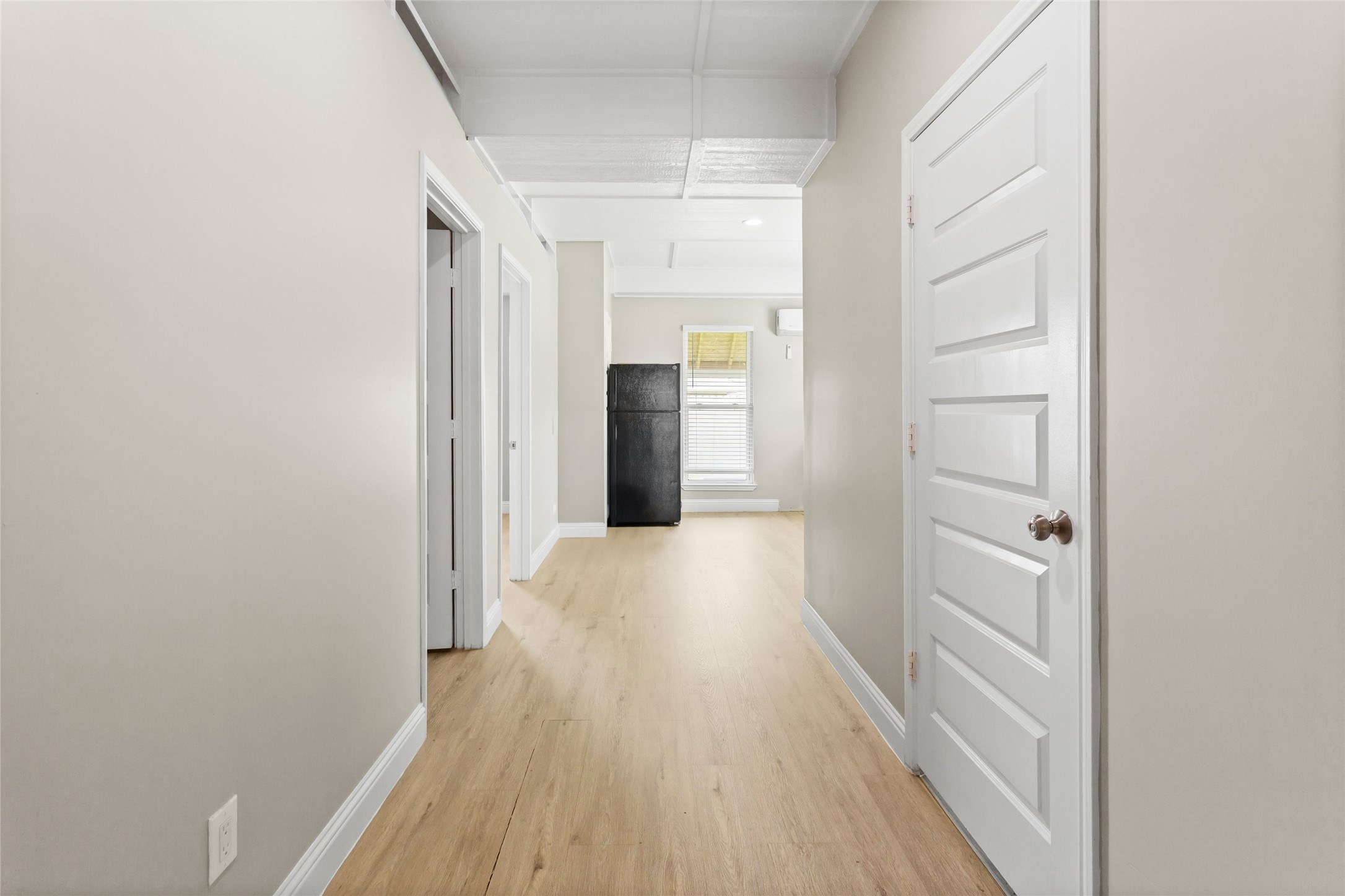 610 South Shady Lane La Porte, TX 77571 - Photo 39 of 50 Bright and airy hallway with light wood laminate floors, neutral walls, and white doors. The space is well-lit with natural light from a window at the end, and features a visible black refrigerator, suggesting proximity to a kitchen area.