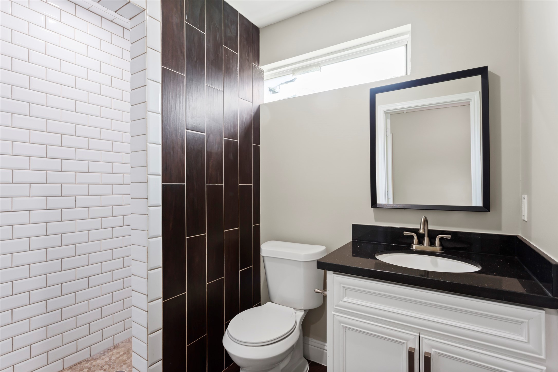610 South Shady Lane La Porte, TX 77571 - Photo 43 of 50 This bathroom features a modern design with a white subway-tiled shower, dark wood accent wall, and a sleek black countertop on a white vanity. A rectangular mirror and a high window provide ample light.