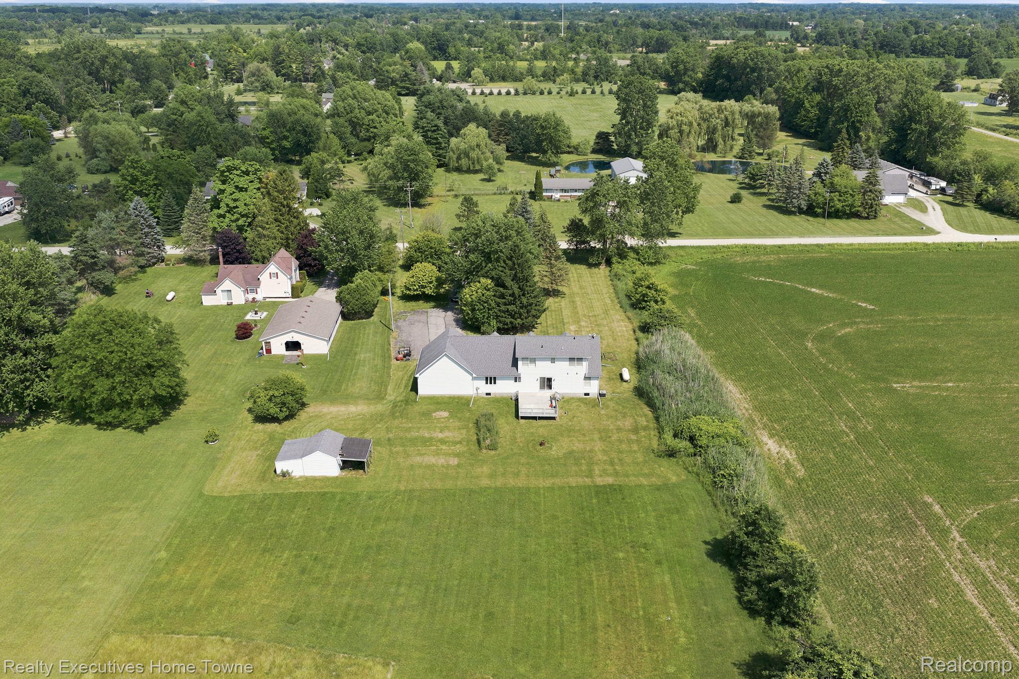 959 Braidwood Road Memphis, MI 48041 - Photo 57 of 58 Expansive property featuring a white two-story residence with a rear deck, a detached garage, a separate shed, and a vast surrounding landscape of green fields and mature trees