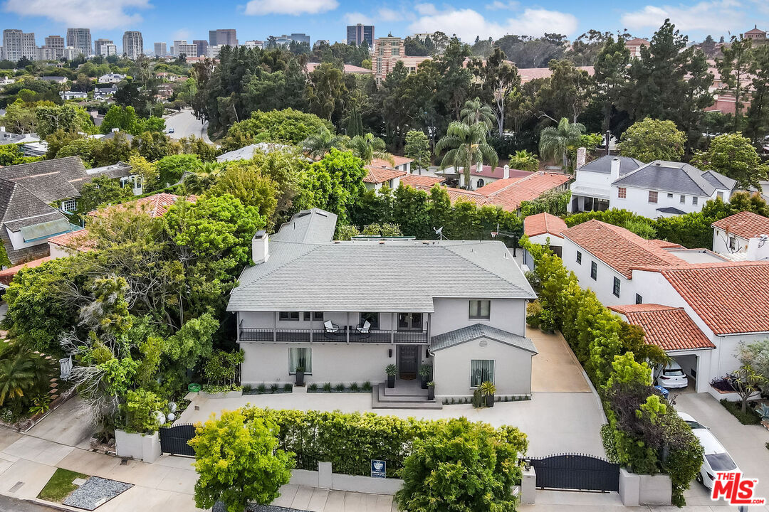  Aerial view of a large, modern home with a lush green yard and palm trees, located in a residential neighborhood. In the background, city skyscrapers and greenery can be seen. The property features a gated entrance, a paved driveway, and a balcony with two chairs. The surrounding homes have red-tiled roofs.