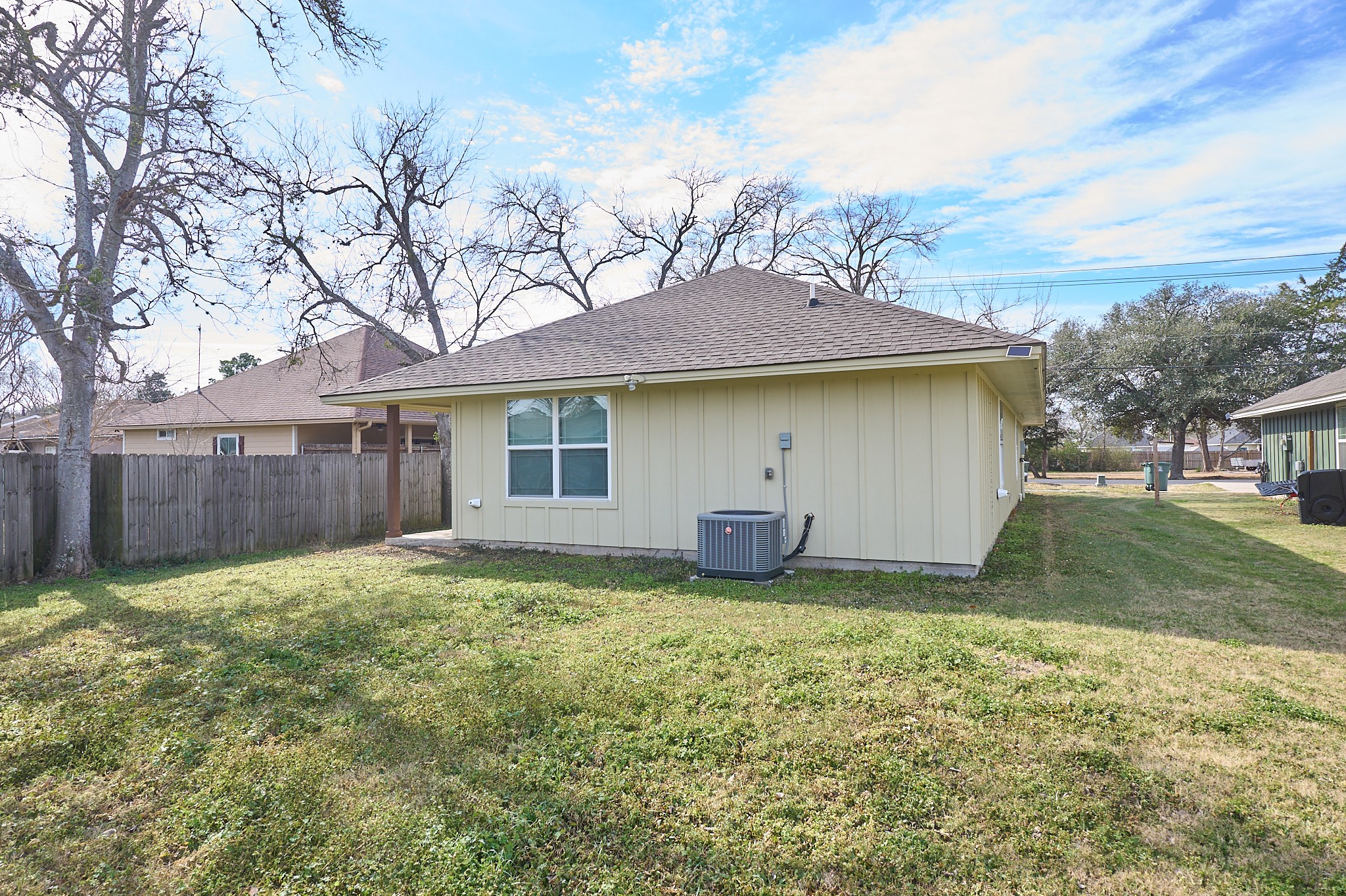1807 Nuches Lane Bryan, TX 77803 - Photo 25 of 27 This photo showcases the backyard of a single-story home with a light yellow exterior. The yard is fenced, featuring a grassy lawn and mature trees, and there's an air conditioning unit visible. It's a spacious outdoor area suitable for various activities.