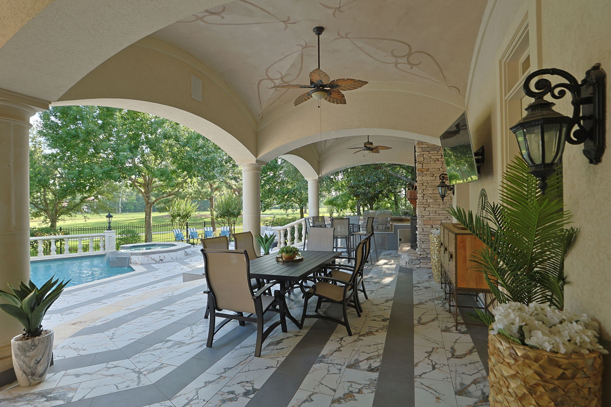 39 Maymont Way Spring, TX 77382 - Photo 37 of 50 This spacious outdoor patio features elegant arches, a dining area, ceiling fans, and a built-in grill. It overlooks a lush garden and a pool, providing a serene and inviting space for relaxation and entertainment.