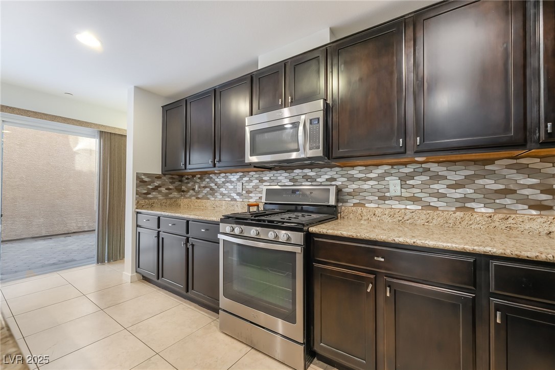 3052 Camino Rico Avenue Henderson, NV 89044 - Photo 17 of 39 Kitchen featuring stainless steel appliances, backsplash, dark brown cabinetry, light tile patterned floors, and light stone countertops