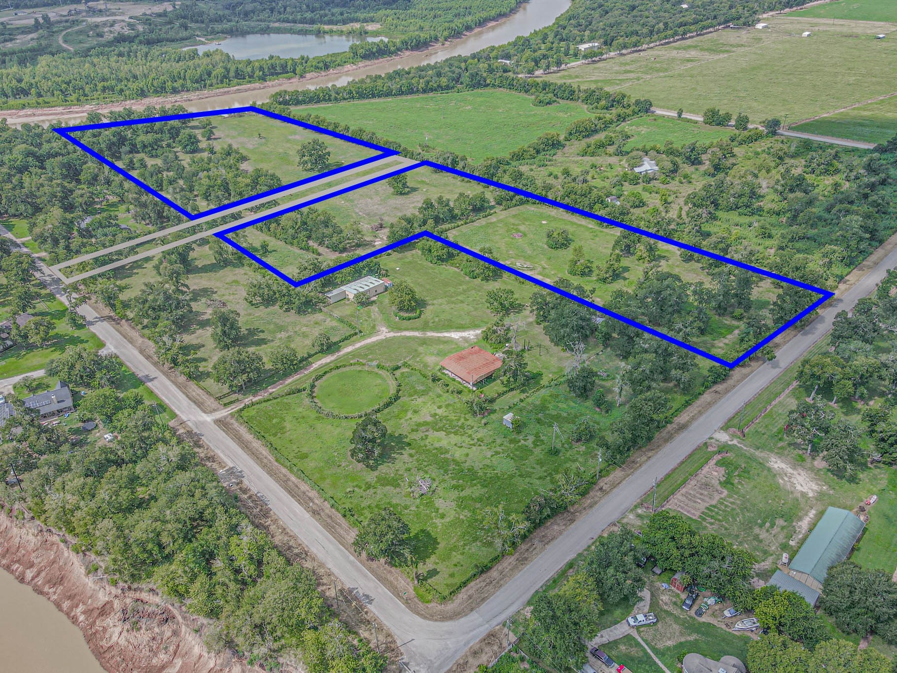This aerial photo shows a spacious rural property outlined in blue, The 6.22 acres for sale is the blue ouline of a large L.  featuring expansive green fields with scattered trees. The land is accessible via nearby roads and is bordered by lush greenery, offering a secluded and serene setting.