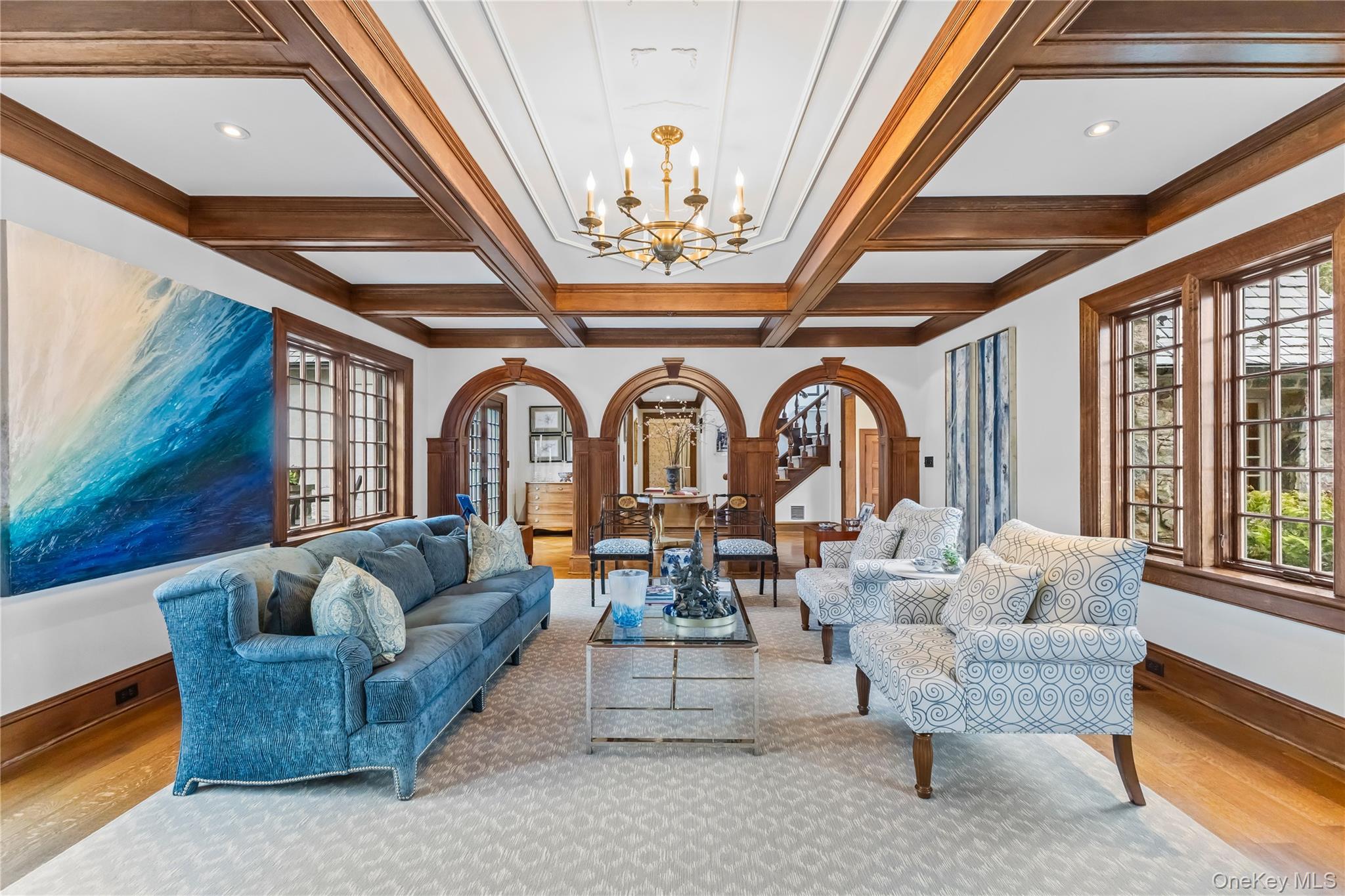 19 Park Drive South Rye, NY 10580 - Photo 10 of 42 Stunning living room showcasing timeless architectural detail with a coffered ceiling, rich wood trim, elegant chandelier, and beautiful arched doorways that create a seamless flow to adjacent spaces. Large windows fill the room with natural light, highlighting the hardwood floors and spacious layout perfect for both everyday living and sophisticated entertaining.
