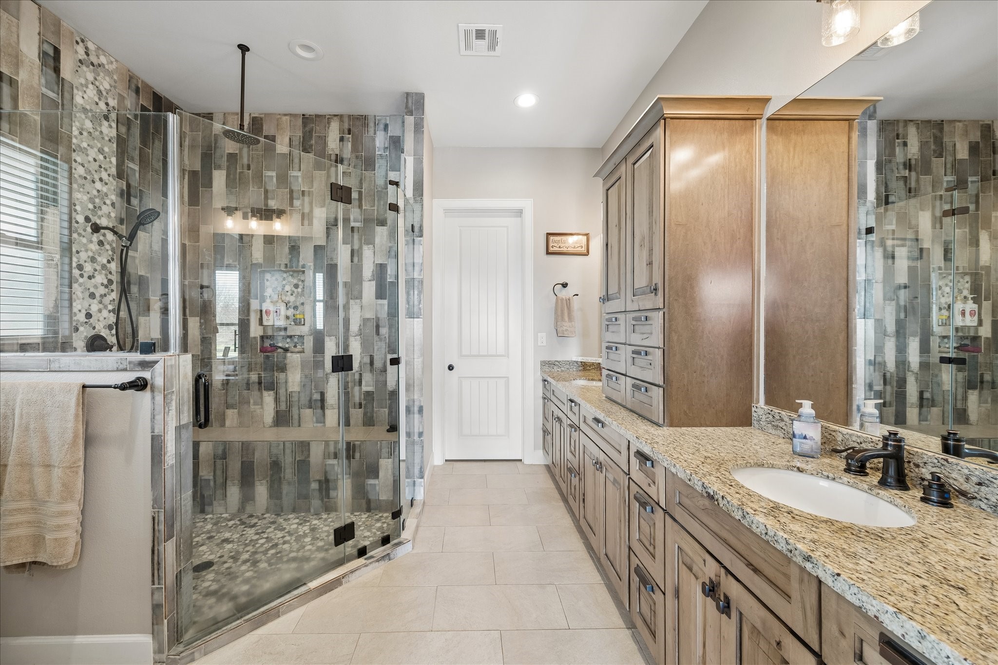 2224 Maler Road Sealy, TX 77474 - Photo 22 of 50 Primary bathroom features a spacious walk-in shower with a modern tile design and clear glass enclosure. It includes a double vanity with granite countertops and ample wooden cabinetry for storage.