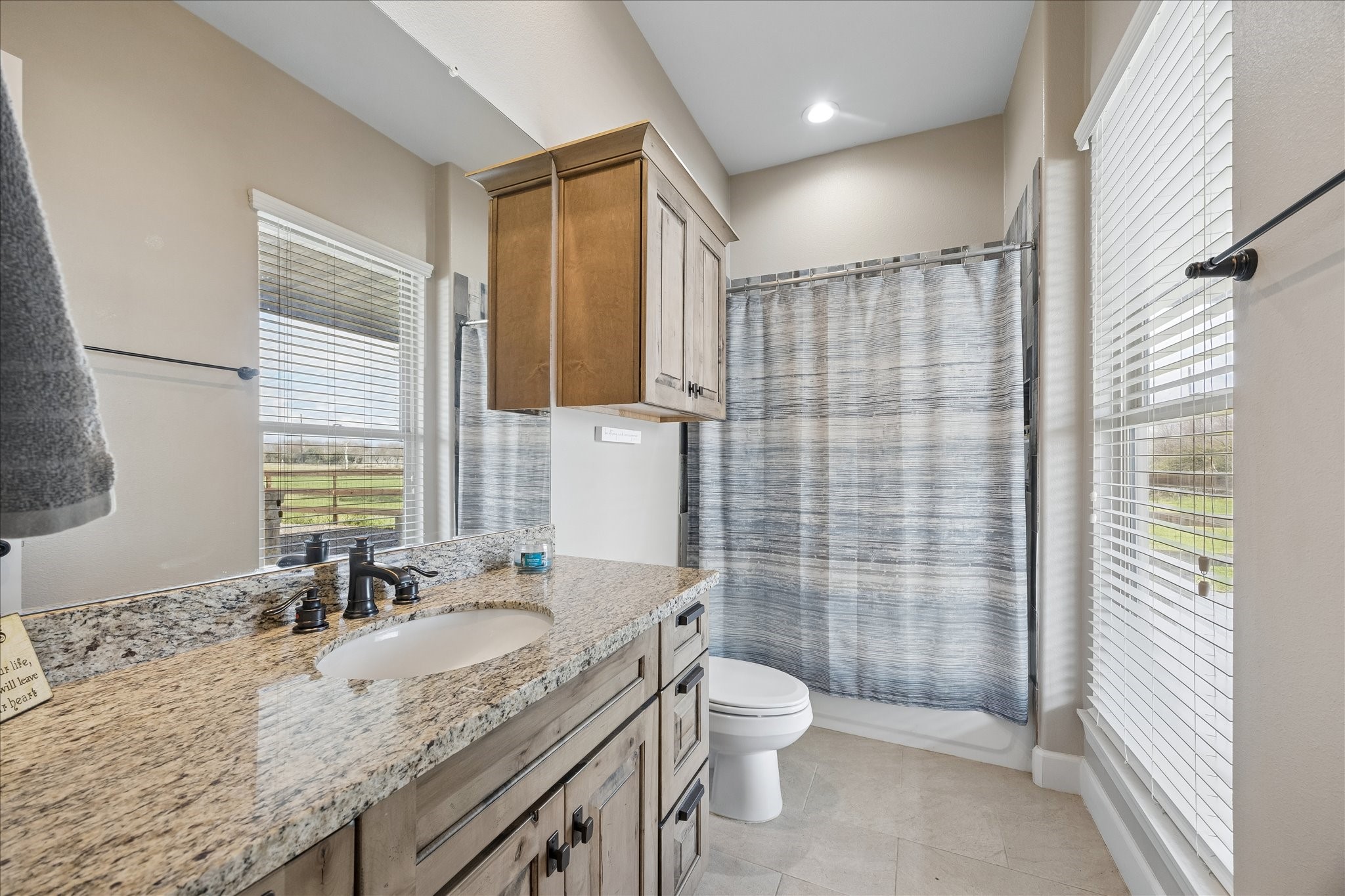 2224 Maler Road Sealy, TX 77474 - Photo 27 of 50 This bathroom features a granite countertop with a single sink, wooden cabinetry, and a shower and large windows for natural light and neutral tiles for a clean, bright look.