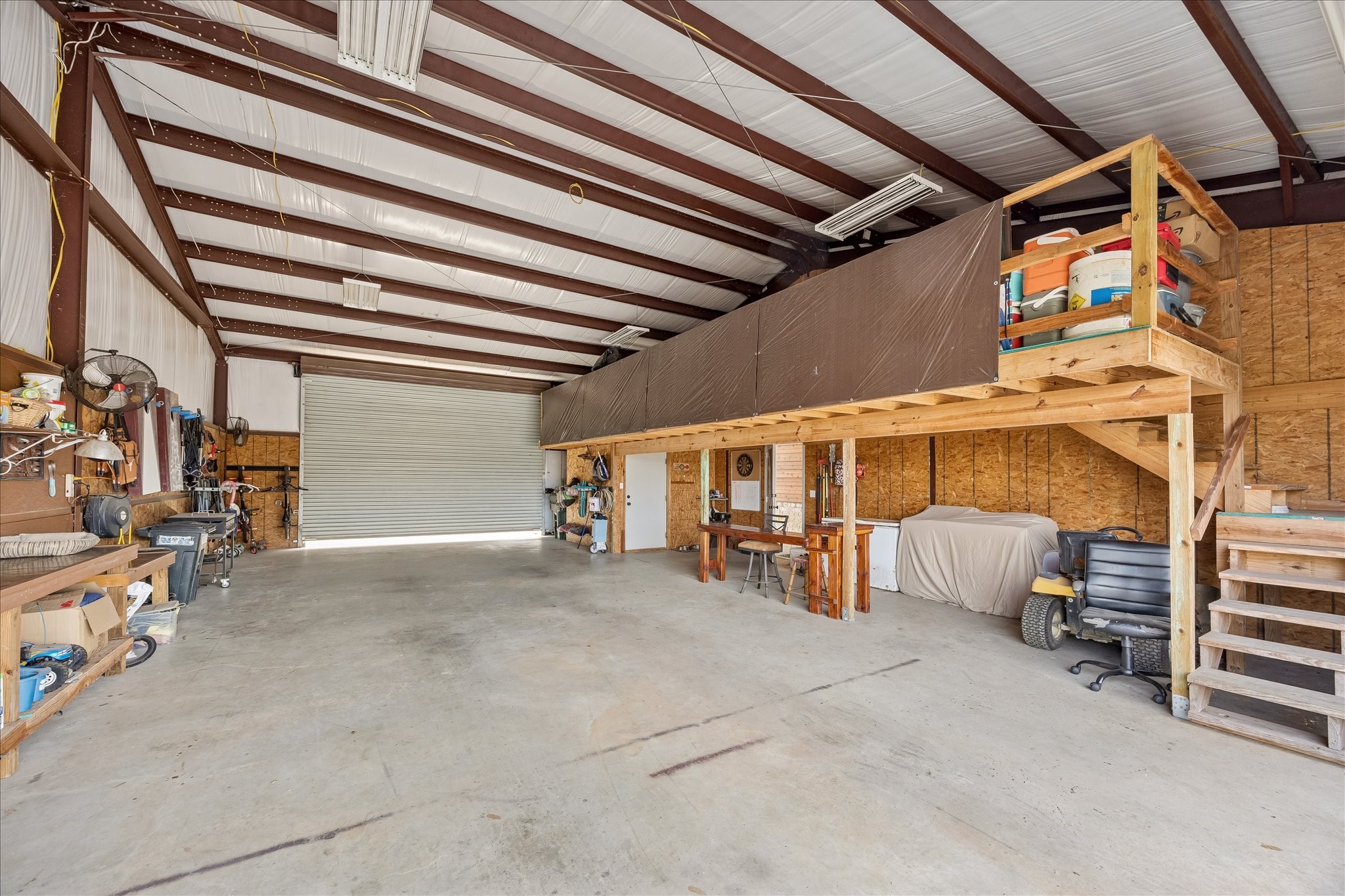 2224 Maler Road Sealy, TX 77474 - Photo 30 of 50 This spacious workshop features a high ceiling with exposed beams, ample storage space, a large garage door, and a mezzanine for additional storage or workspace. Ideal for projects or hobbies.