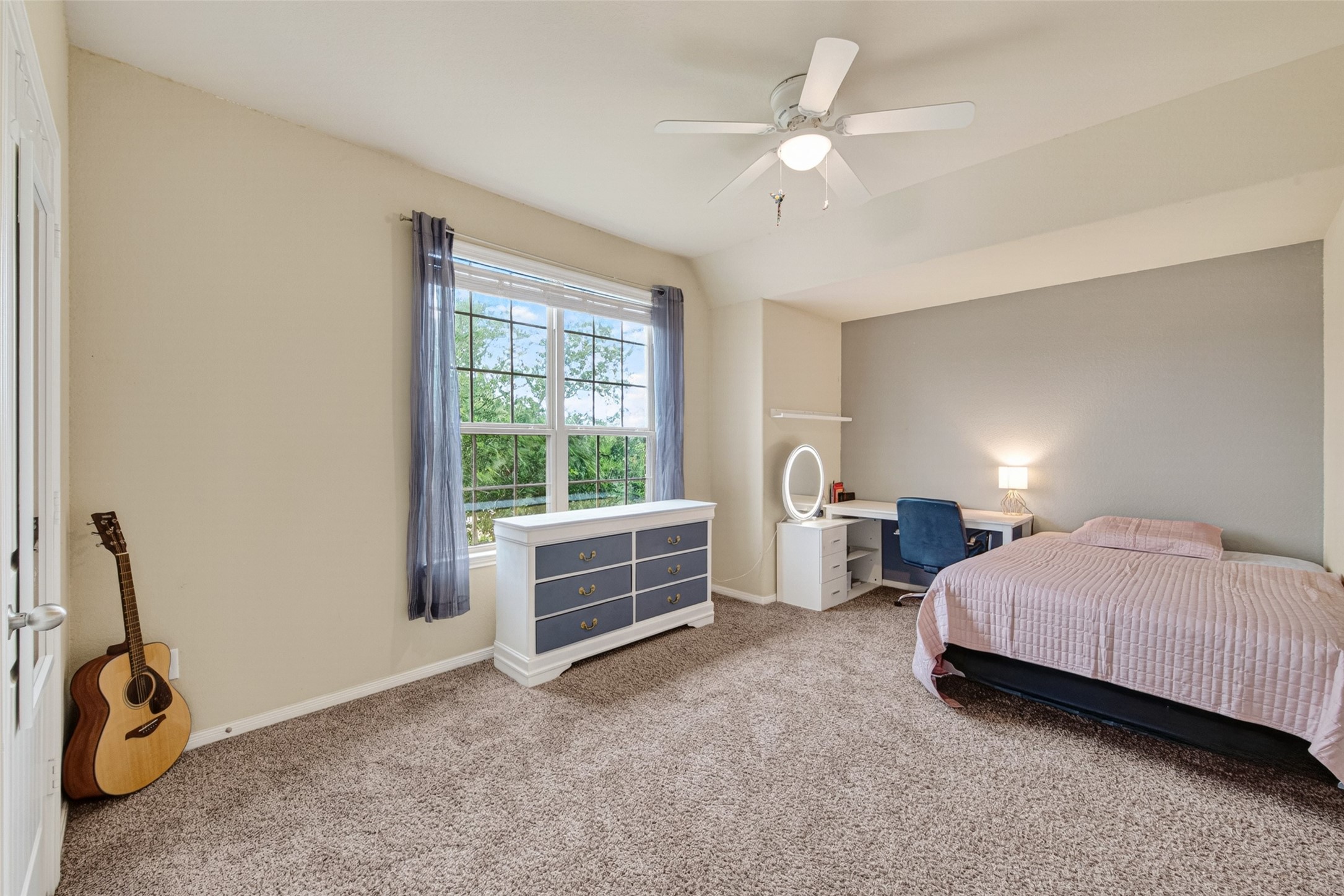 25611 Ellis Ridge Court Katy, TX 77494 - Photo 28 of 49 This bright bedroom features a large window with sheer curtains, beige walls, and carpeted flooring. It includes a ceiling fan, a cozy bed, a desk with a chair, and a dresser with modern accents. An acoustic guitar adds a touch of personality.