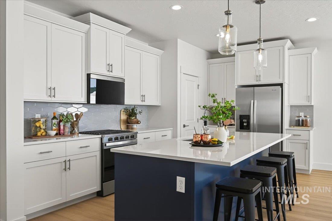 14880 Wing Spread Drive Caldwell, ID 83607 - Photo 4 of 12 Contemporary kitchen featuring an expansive center island with seating, white shaker-style cabinetry, stainless steel appliances, a hexagonal tile backsplash, and light wood-finish flooring