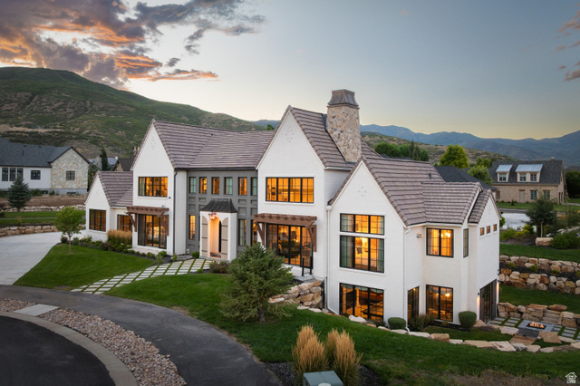 558 West Rheinfalls Circle Midway, UT 84049 - Photo 2 of 61 Modern farmhouse estate featuring a striking white exterior with multiple peaked rooflines and a tall stone chimney, expansive multi-pane windows with warm interior lighting, manicured lawn with ornamental grasses, and a circular driveway with permeable p