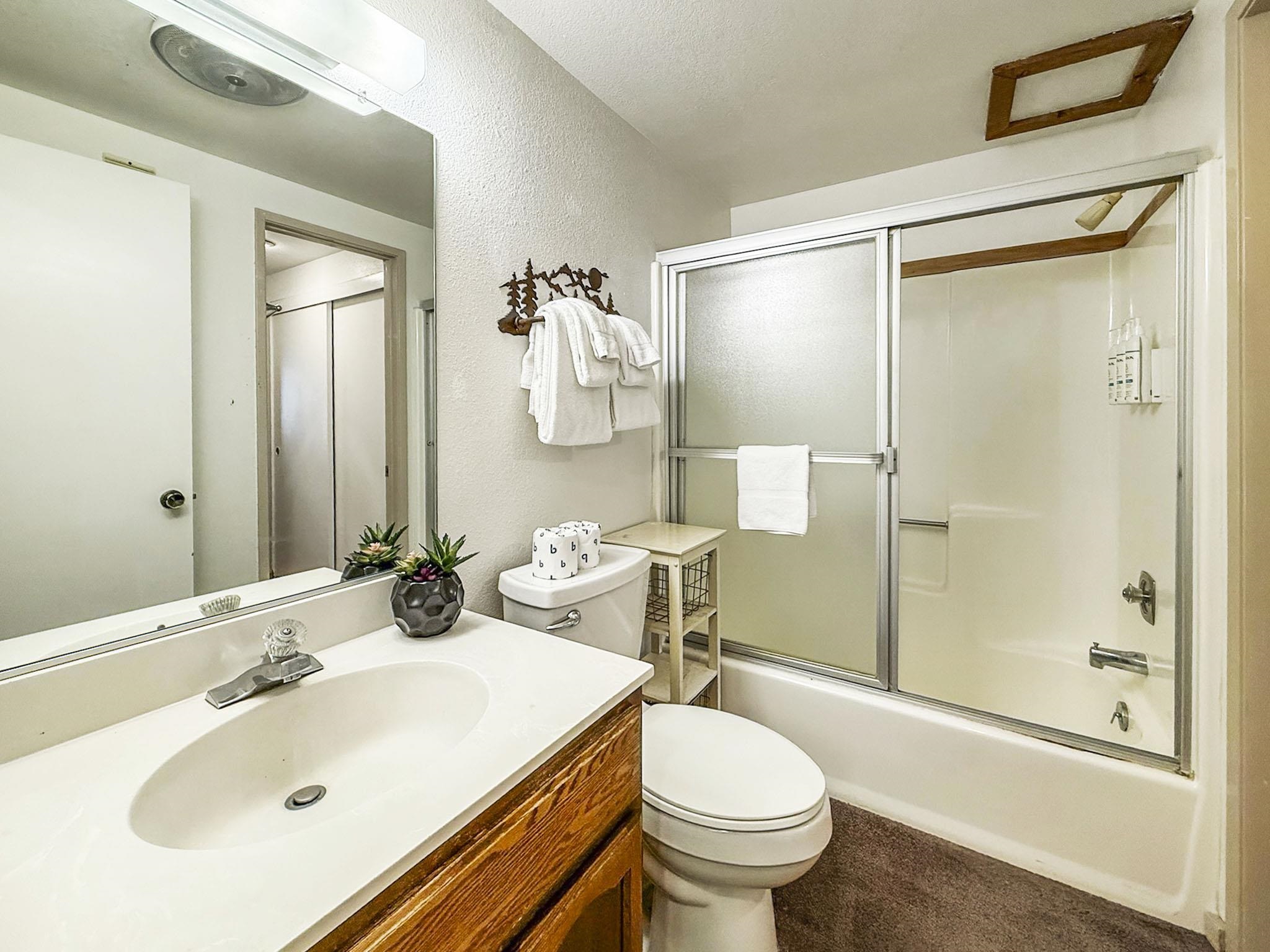 153 Lake Mary Road, Unit 114 Mammoth Lakes, CA 93546 - Photo 12 of 22 Bathroom featuring a shower-tub combination with sliding frosted glass doors, a vanity with a white countertop and integrated sink, a wood-finish cabinet base, and a wall-mounted mirror