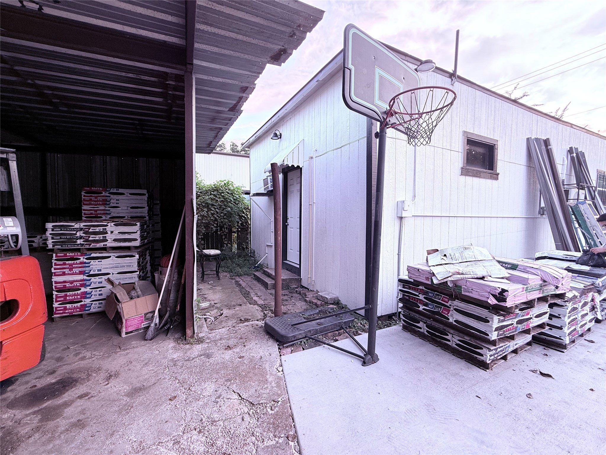 6325 Cullen Boulevard Houston, TX 77021 - Photo 14 of 18 This photo shows a backyard area with a covered storage space and a white exterior building. There are stacks of materials, possibly construction supplies, and a basketball hoop. The space appears functional for storage or projects.