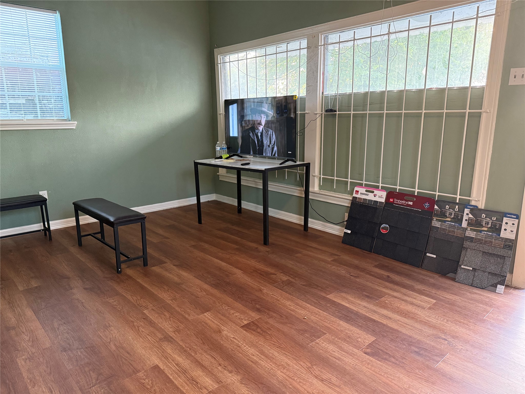 6325 Cullen Boulevard Houston, TX 77021 - Photo 17 of 18 This room features wood flooring and large windows with security bars, allowing for natural light. It's currently set up with a small table and bench, and has samples of roofing materials displayed, offering potential for varied use.