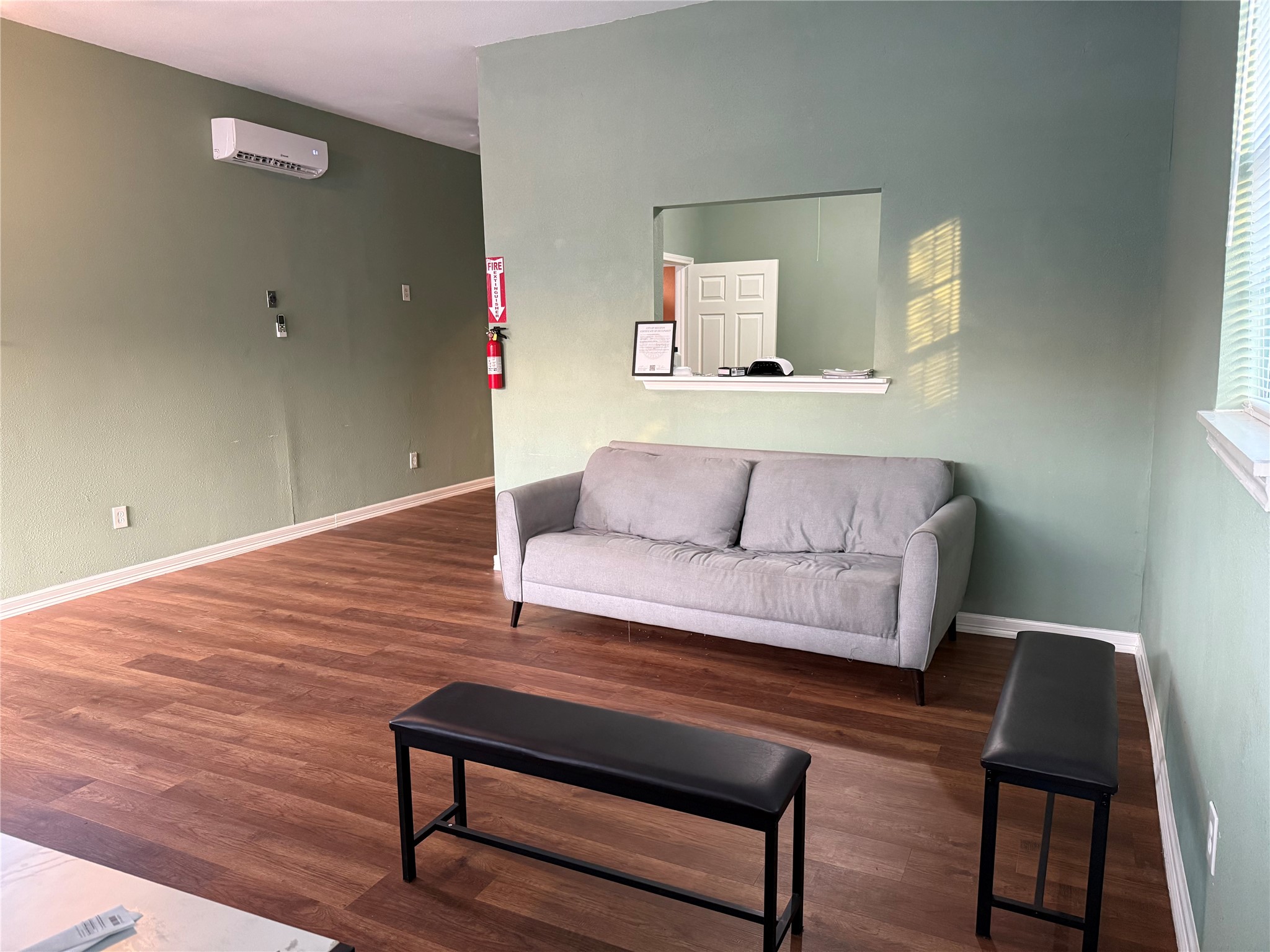 6325 Cullen Boulevard Houston, TX 77021 - Photo 2 of 18 This room features a cozy gray sofa against a soft green wall, with a bench and wood flooring. It has a wall-mounted air conditioner and a pass-through window to an adjacent space, creating an open, airy feel.