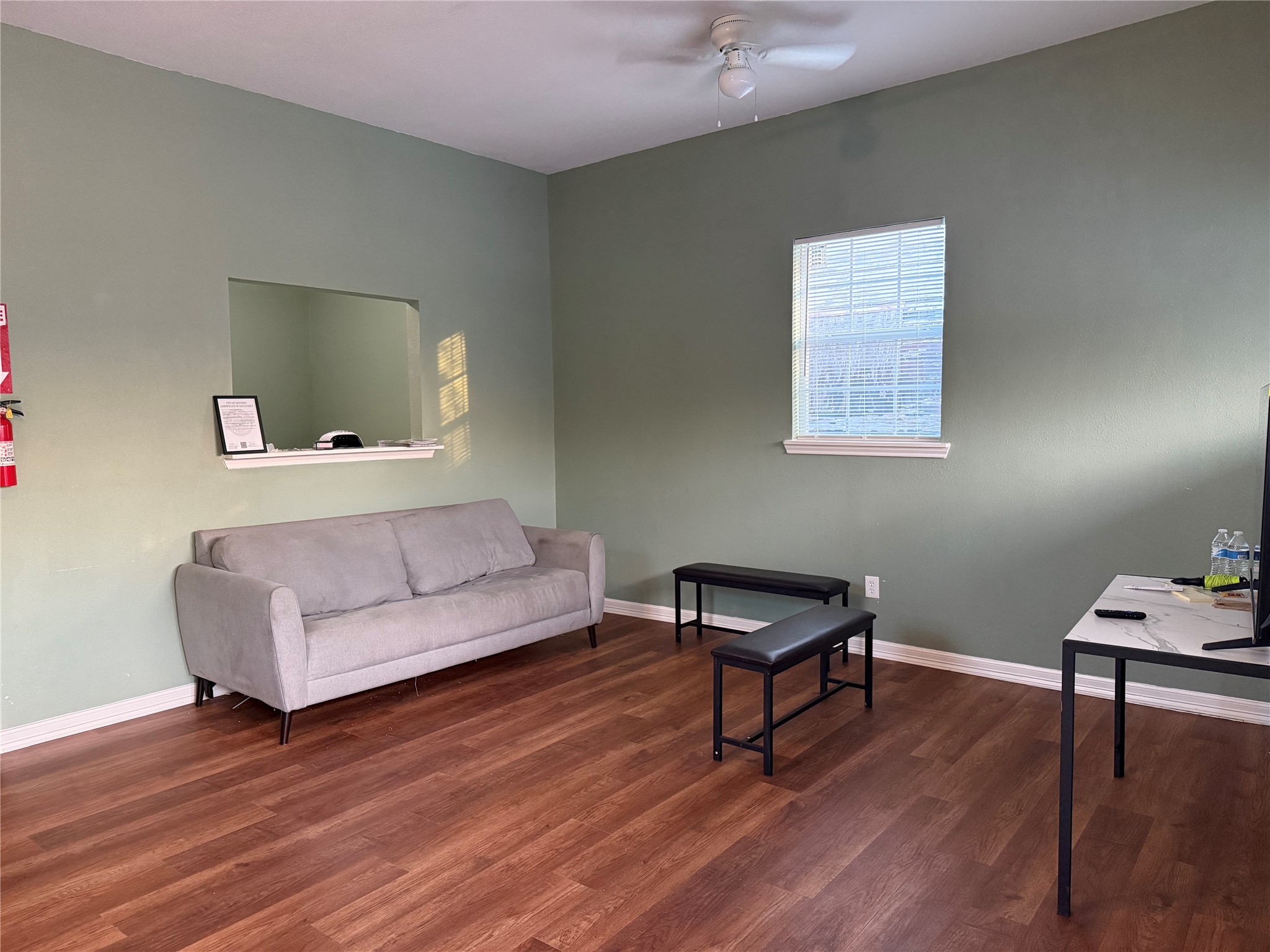 6325 Cullen Boulevard Houston, TX 77021 - Photo 3 of 18 This room features a cozy gray sofa against a soft green wall, complemented by wood-style flooring. A small window allows natural light, and there's a simple ceiling fan above. The space is minimally furnished with a TV and a small table, creating a relaxed and functional living area.