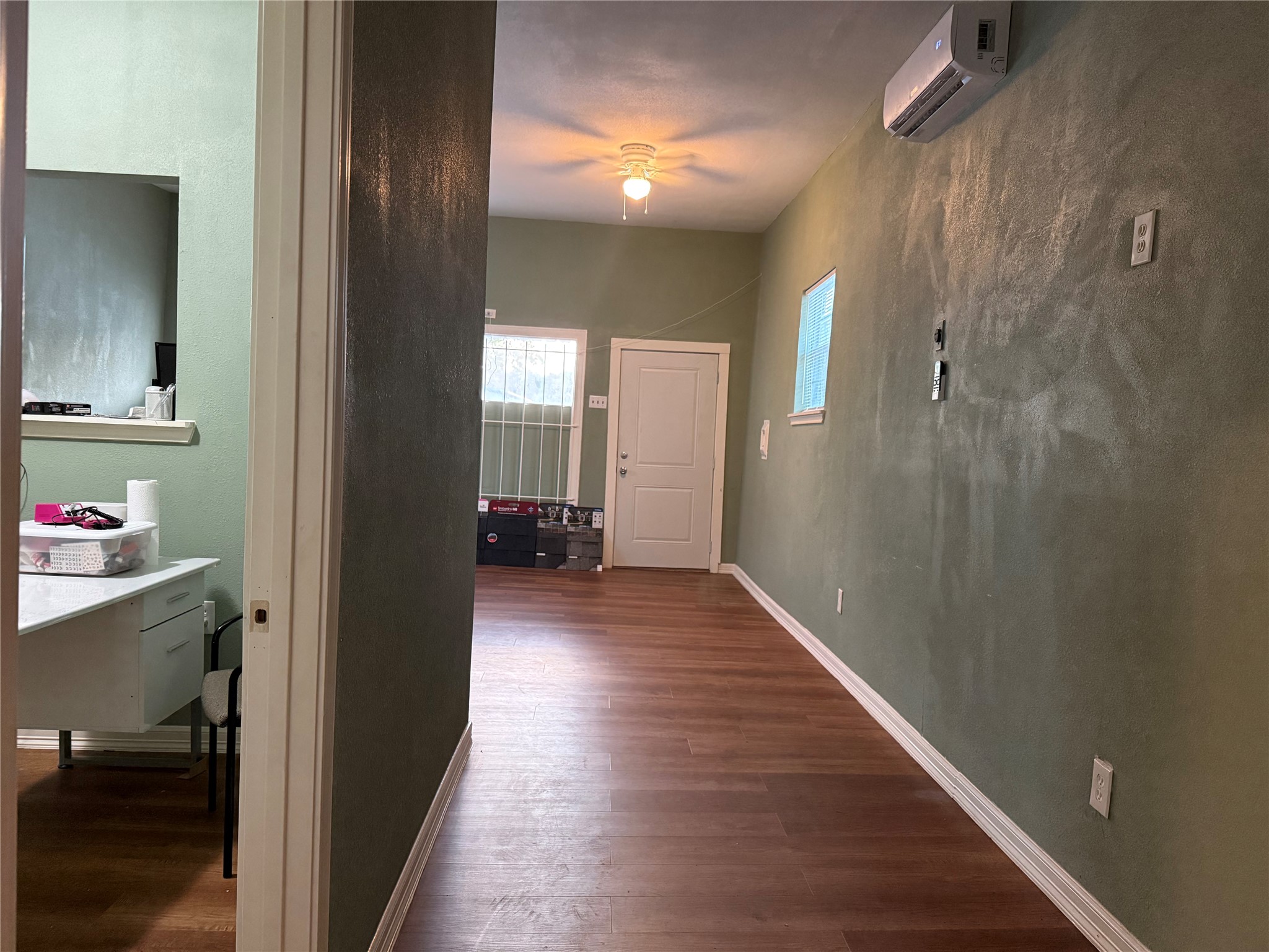 6325 Cullen Boulevard Houston, TX 77021 - Photo 5 of 18 This photo shows a narrow room with light green walls and wood flooring. It has a wall-mounted air conditioner, a small window, and a ceiling light. Visible is an adjacent room with a desk area, and a door leading outside.
