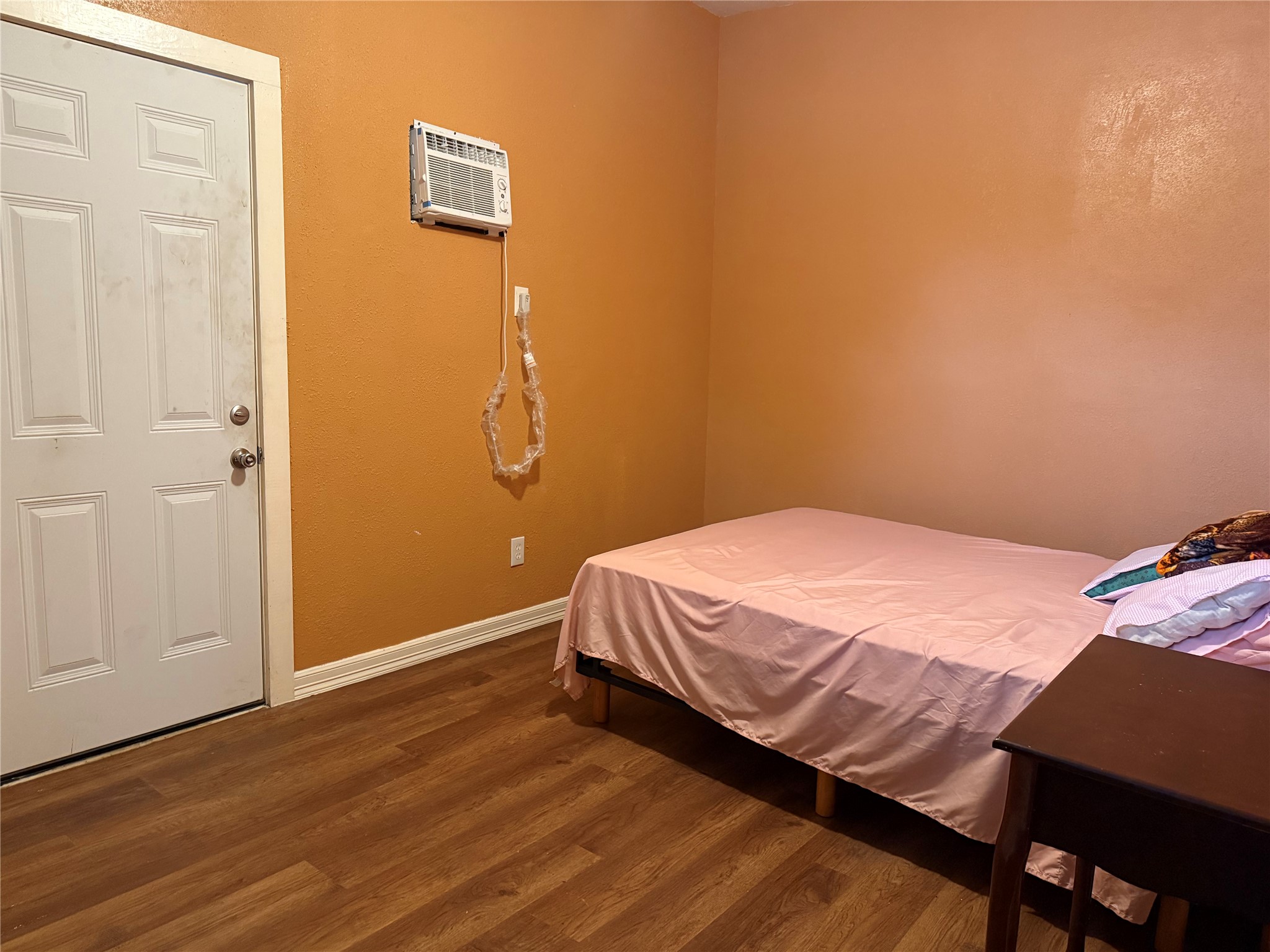 6325 Cullen Boulevard Houston, TX 77021 - Photo 8 of 18 This cozy bedroom features warm orange walls, wood flooring, and a compact layout with a bed and small table. It includes a wall-mounted air conditioner and a door with direct outdoor access.
