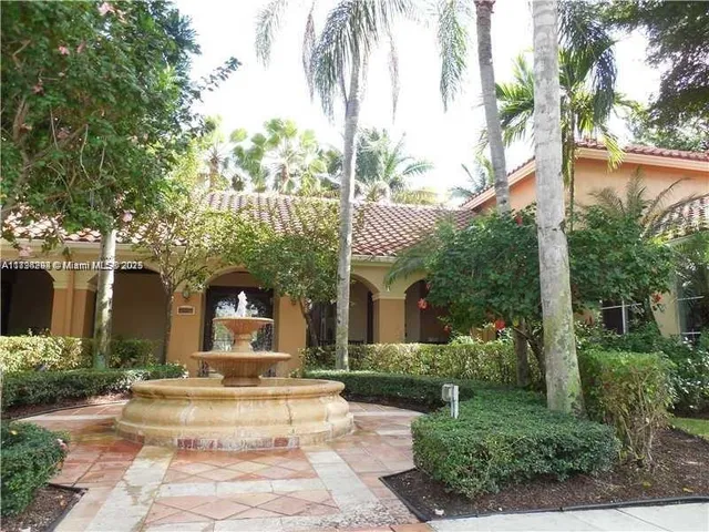 a view of a white house with a fountain plants and large tree