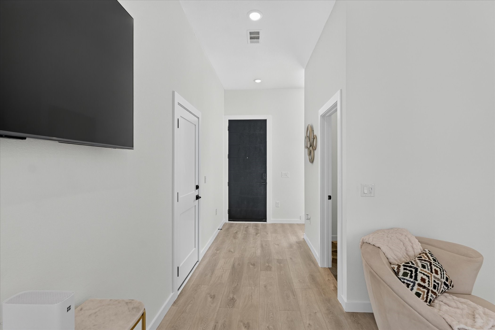 2000 Riverside Farms Road, Unit 8101 Austin, TX 78741 - Photo 8 of 41 This photo showcases a modern, bright hallway with light wood flooring and white walls. It features a mounted TV, a black door at the end, and a cozy chair with decorative pillows, creating a welcoming entryway.
