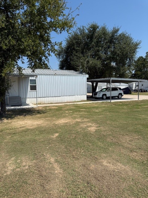 22786 Cuttler Road New Caney, TX 77357 - Photo 3 of 12 This photo shows a modest mobile home with a metal exterior, surrounded by a grassy yard. It features a covered carport and is partially shaded by large trees, suggesting a quiet and simple living space.