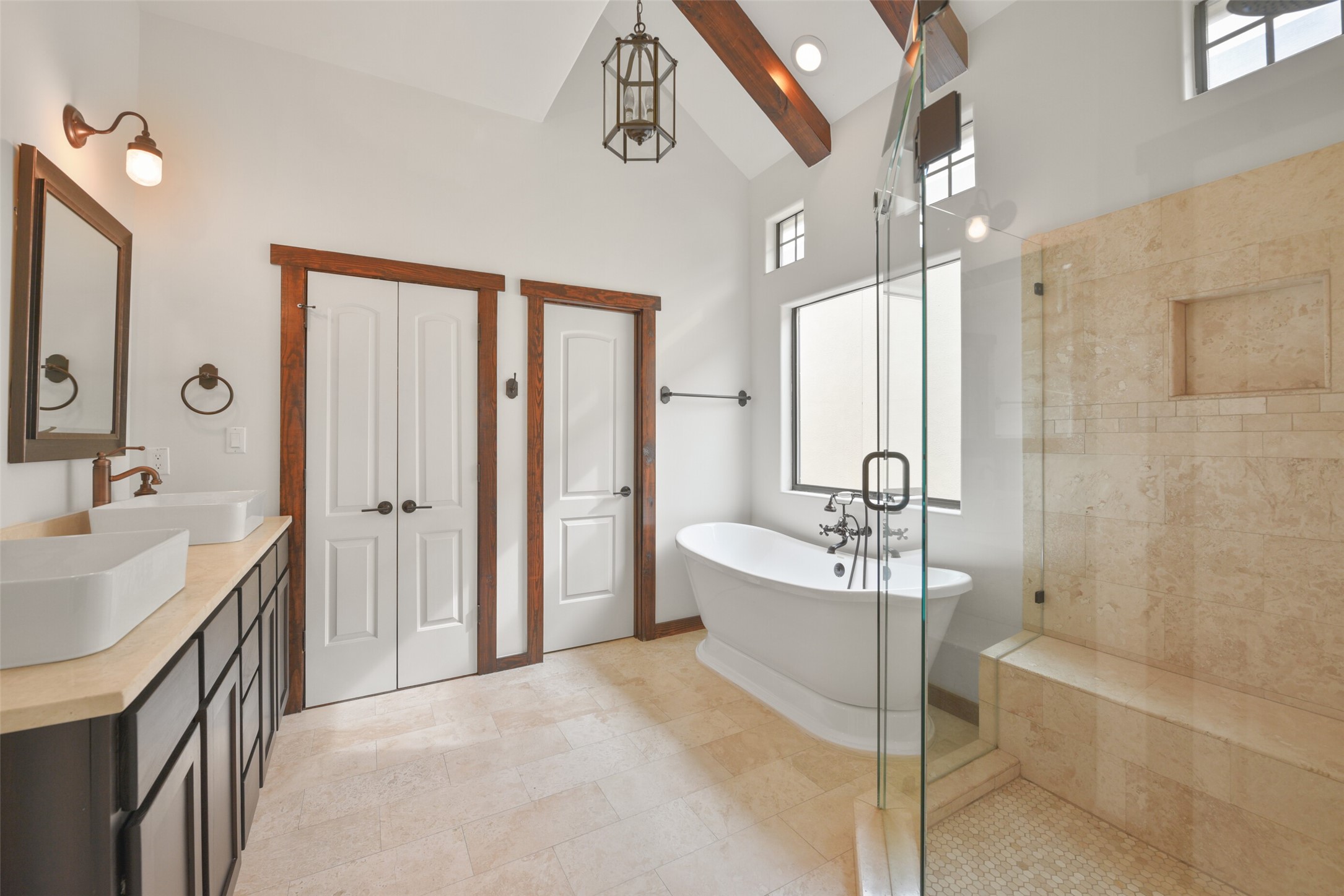 2601 Bevis Street, Unit B Houston, TX 77008 - Photo 15 of 25 Luxurious Primary Bathroom featuring travertine floors and countertop, a separate shower, freestanding tub, large vanity w/ 2 sinks, & ample storage. On the right is a separate water closet. High ceilings with rustic wood beams enhance the space w/ decorative light fixture. French doors lead to a large walk-in closet w/ built-in floor-length mirror and shelving for all your storage needs.