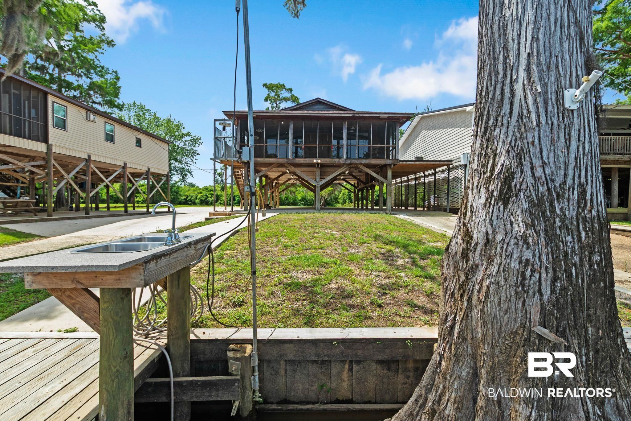 8153 Bryant's Lndg Road Stockton, AL 36579 - Photo 57 of 87 Waterfront property featuring a raised structure with a screened porch, a ground-level concrete patio, an outdoor sink station, an adjacent boat dock, and a large mature tree