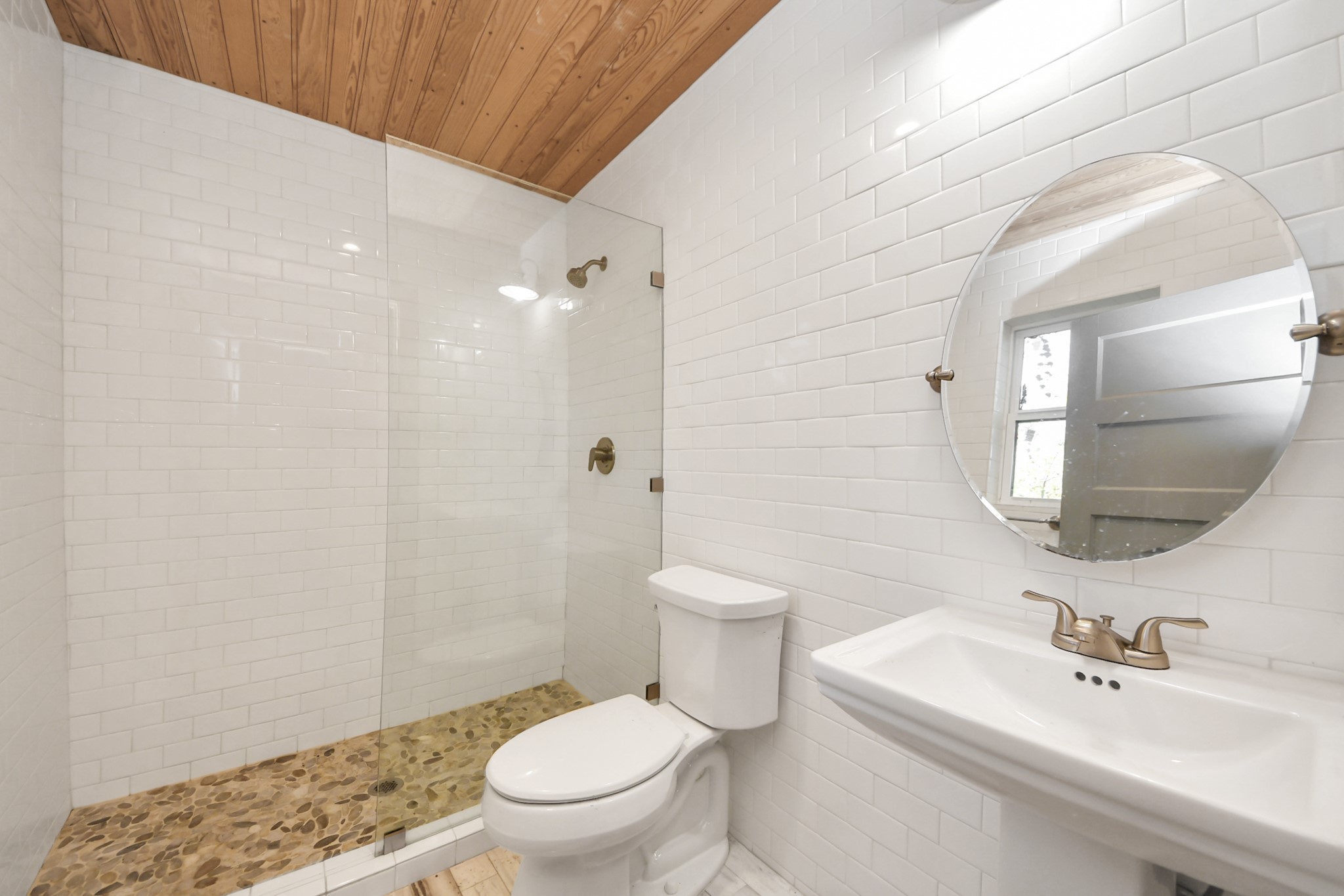 1202 Archer Street Houston, TX 77009 - Photo 42 of 46 This bathroom features a clean, modern design with white subway tile walls and a wood-paneled ceiling. It includes a glass-enclosed shower with pebble flooring, a sleek pedestal sink, and a round mirror. Natural light enters through a small window, enhancing the serene atmosphere.