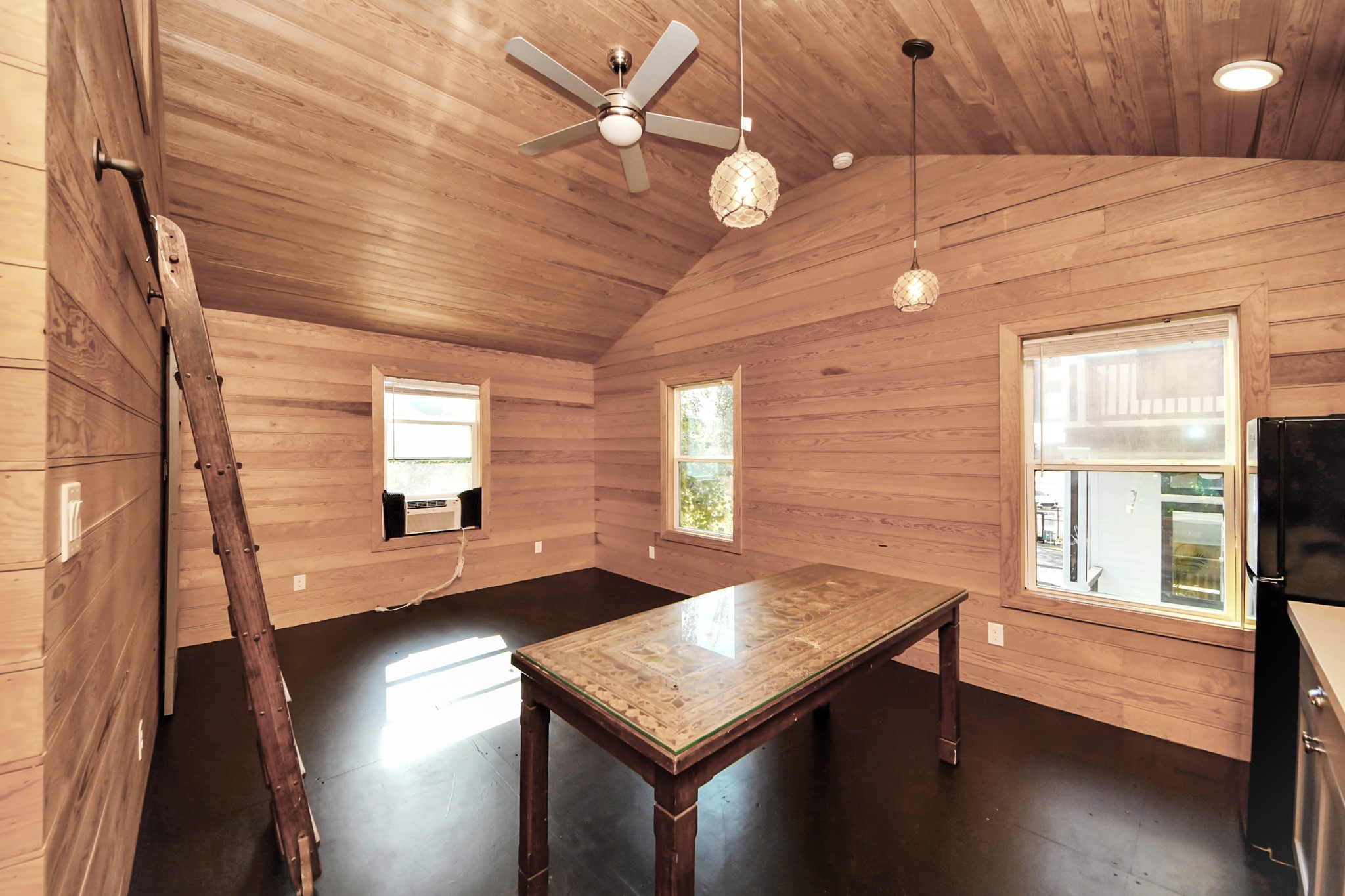 1202 Archer Street Houston, TX 77009 - Photo 45 of 46 This rustic room showcases wood-paneled interiors with vaulted ceilings, a ceiling fan, and pendant lighting. A large central table anchors the space, while two windows fill it with natural light. An air conditioning unit adds comfort, and a ladder hints at potential loft access, combining functionality with rustic charm.