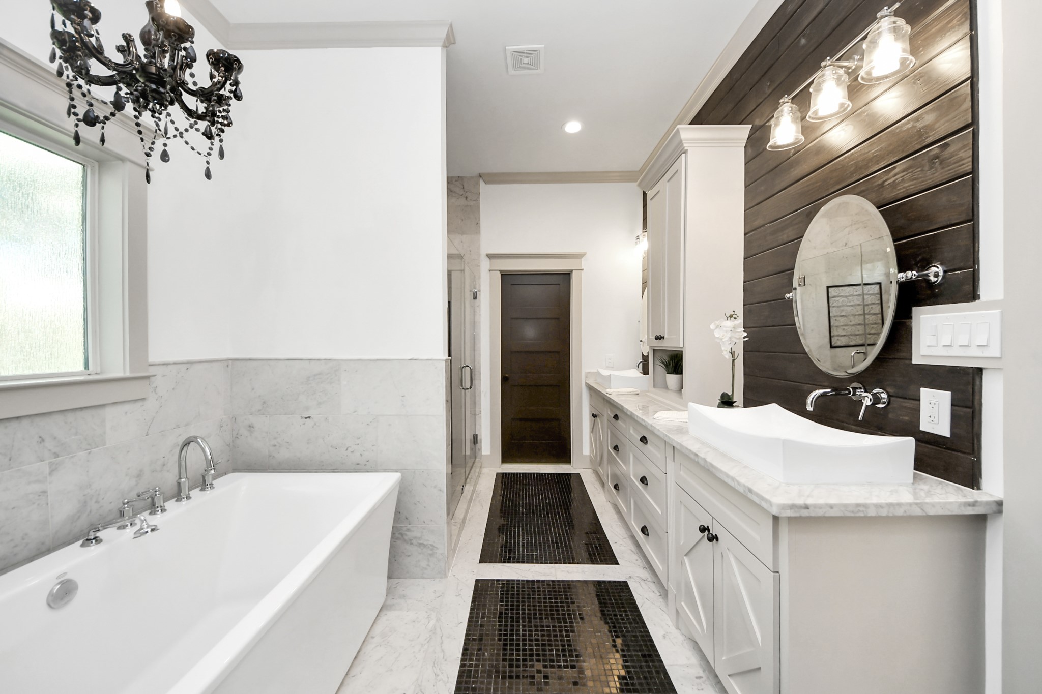 1202 Archer Street Houston, TX 77009 - Photo 8 of 46 This elegant bathroom showcases a luxurious freestanding bathtub, dual sinks set into a marble countertop, and a striking wood-accent wall. Sunlight and modern lighting, along with a sophisticated chandelier, create a bright, serene, and spa-like atmosphere.
