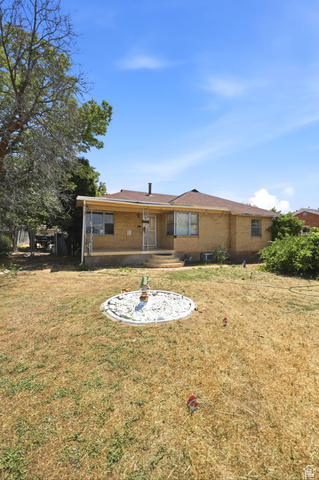 5480 South 2000th Street West Roy, UT 84067 - Photo 6 of 36 Brick exterior with a covered porch, featuring a brown shingle roof, mature trees, and a decorative garden bed in the foreground