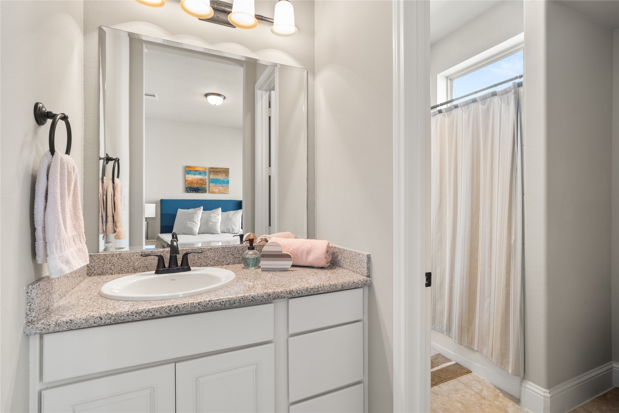 19813 Cullen Ridge Drive Porter, TX 77365 - Photo 35 of 44 This bathroom features a granite countertop with a single sink and black faucet, complemented by a large mirror and modern lighting. Neutral walls and a shower with a curtain create a clean, inviting atmosphere. A glimpse of a bedroom with a blue headboard is visible through the doorway.