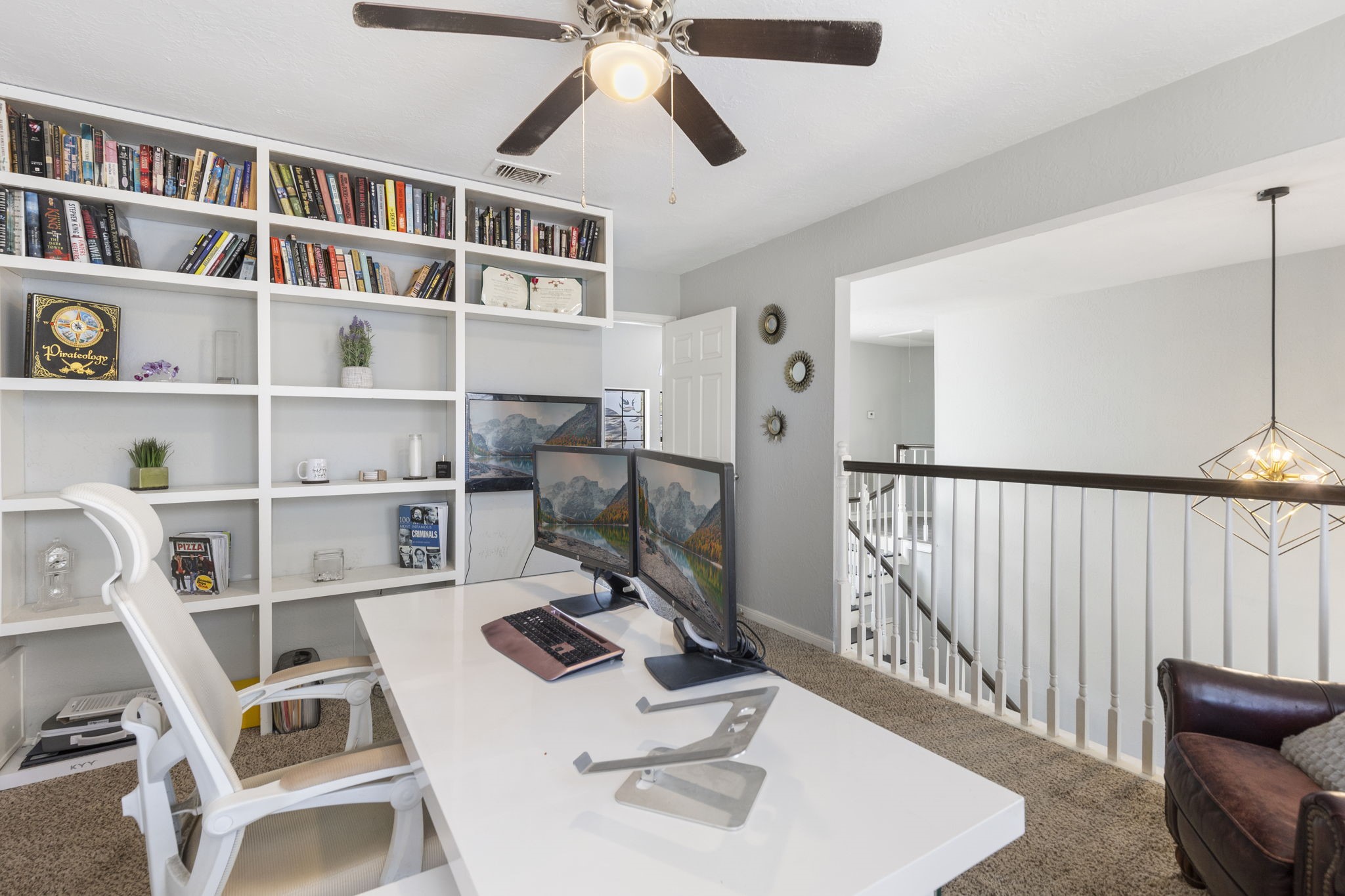 2110 Paso Rello Drive Houston, TX 77077 - Photo 23 of 41 This photo showcases a bright home office with built-in bookshelves and a modern desk setup. It features a ceiling fan, neutral walls, and carpeted flooring. The space is open to a hallway with a railing, enhancing the airy feel.