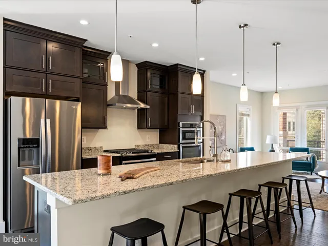 a kitchen with stainless steel appliances granite countertop a sink a refrigerator a kitchen island a stove and chairs with wooden floor