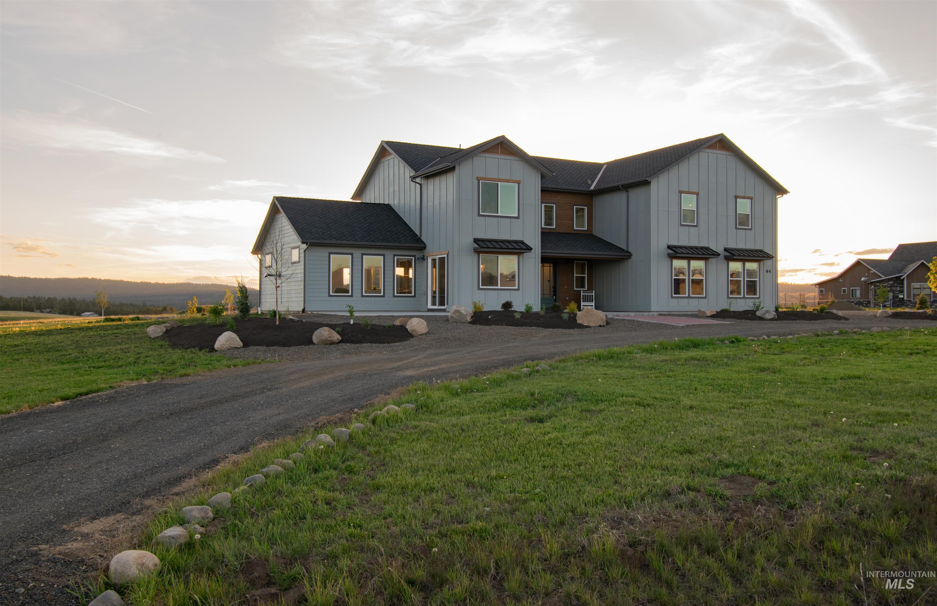 Contemporary farmhouse exterior featuring light gray vertical siding, dark window trim, dark roof, multiple windows, and a gravel driveway