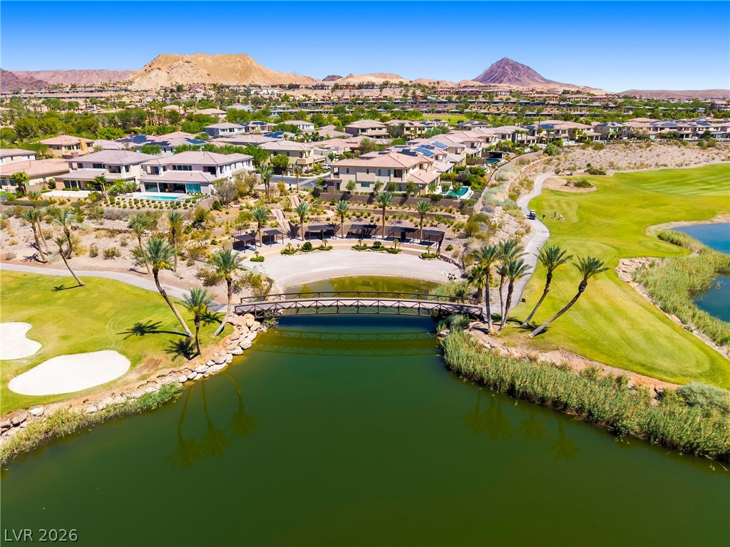 45 Garibaldi Way Henderson, NV 89011 - Photo 56 of 74 An aerial panorama reveals a golf course community with homes nestled among hills, featuring a scenic bridge over a pond.