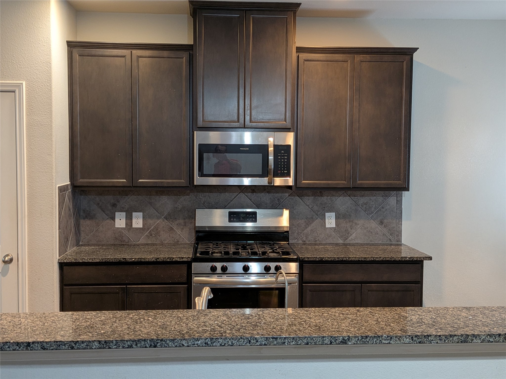 15730 Joe Dimaggio Street Splendora, TX 77372 - Photo 8 of 20 This kitchen features modern dark wood cabinetry, stainless steel appliances including a microwave and gas stove, and a sleek granite countertop with a matching backsplash.