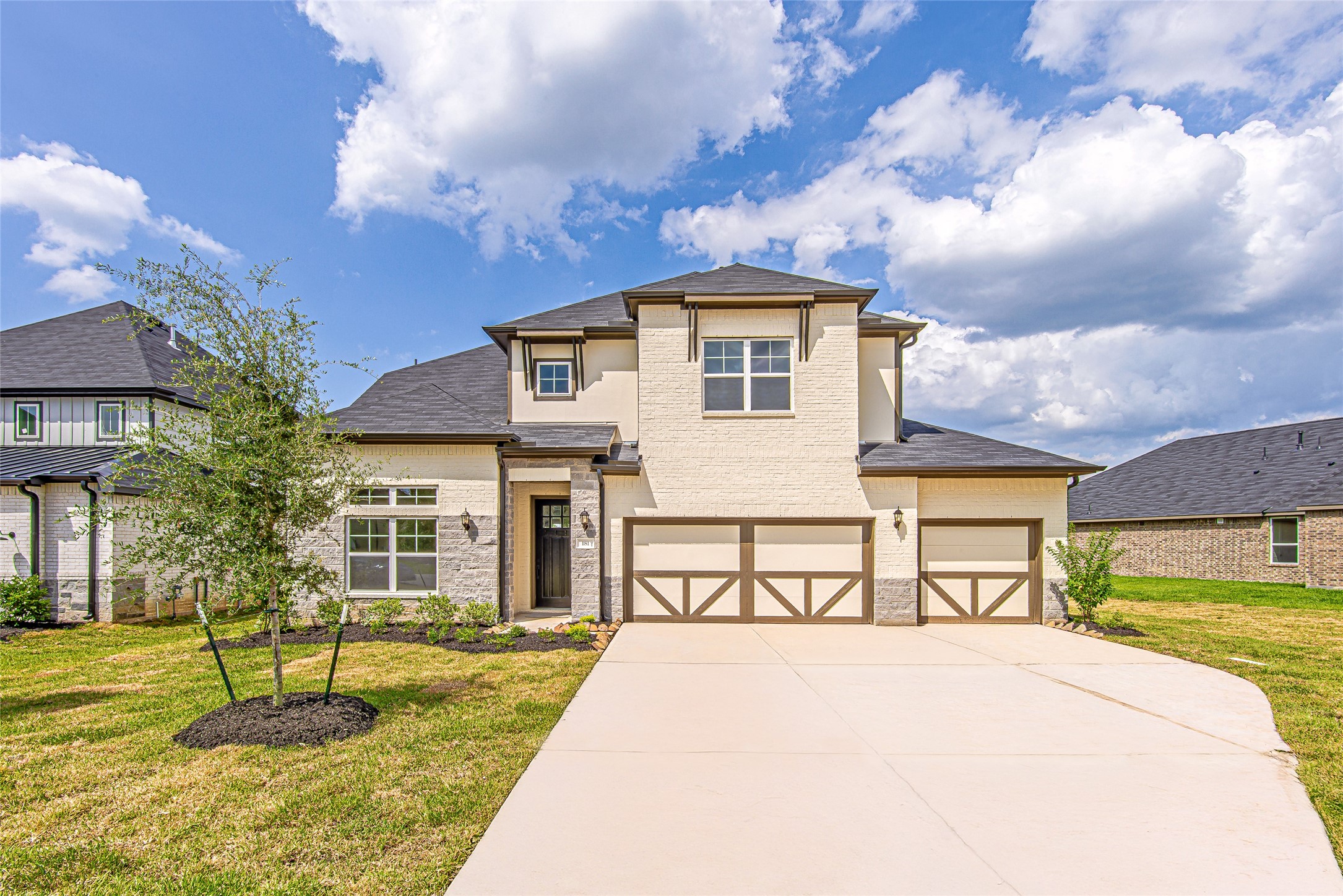 A striking exterior view of the home showcases its elegant modern farmhouse design, featuring contrasting brick and stonework, a deep driveway, and a 3-car garage with crossbuck accents. Manicured landscaping and bright skies frame this eye-catching curb appeal.